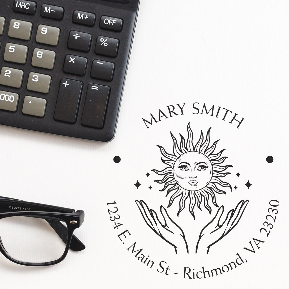 Black custom stamp seal with sun face logo, Mary Smith text, and address on white paper beside a calculator and glasses.