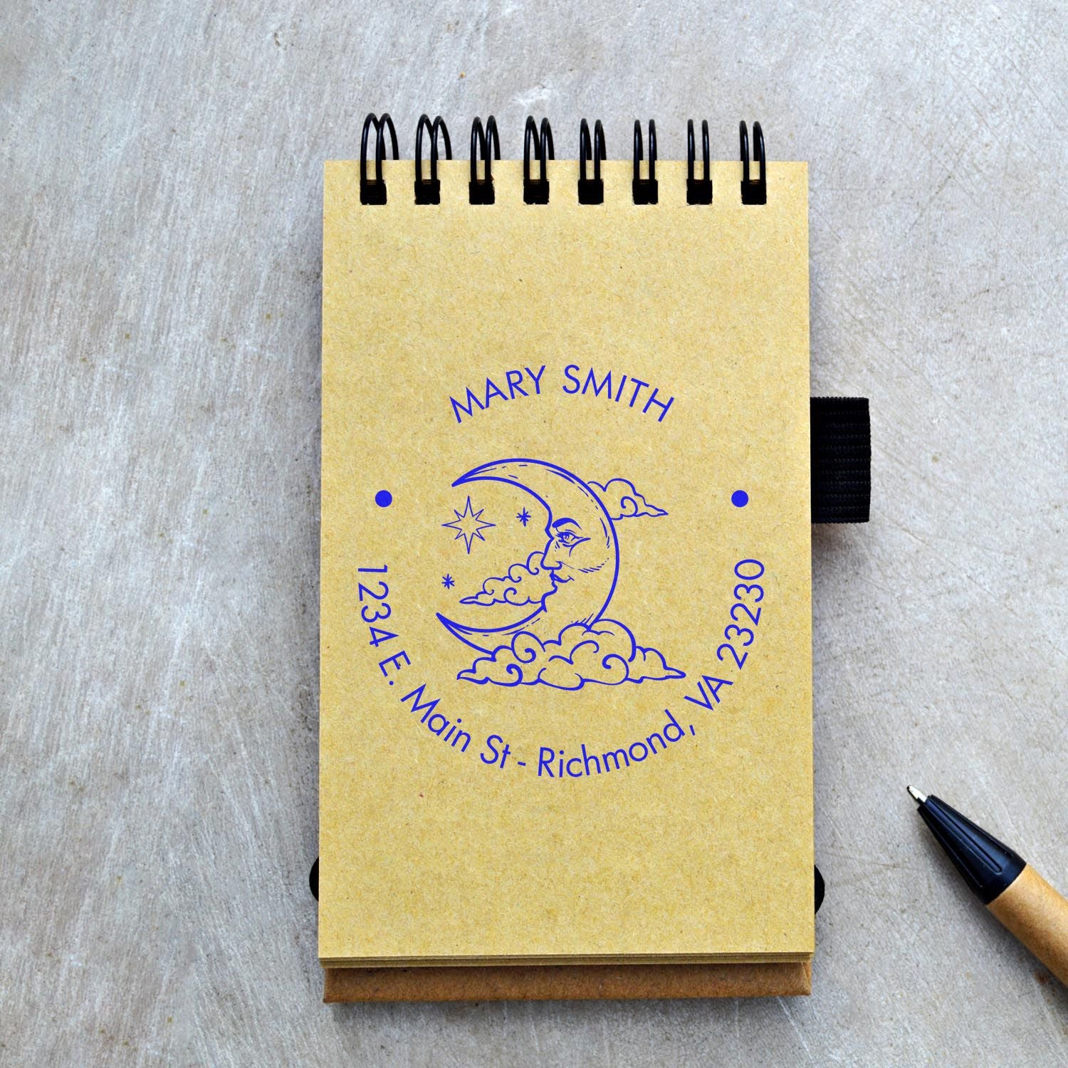 Yellow spiral notebook with blue custom stamp seal, moon and stars design, black binding, and black pen on gray tabletop