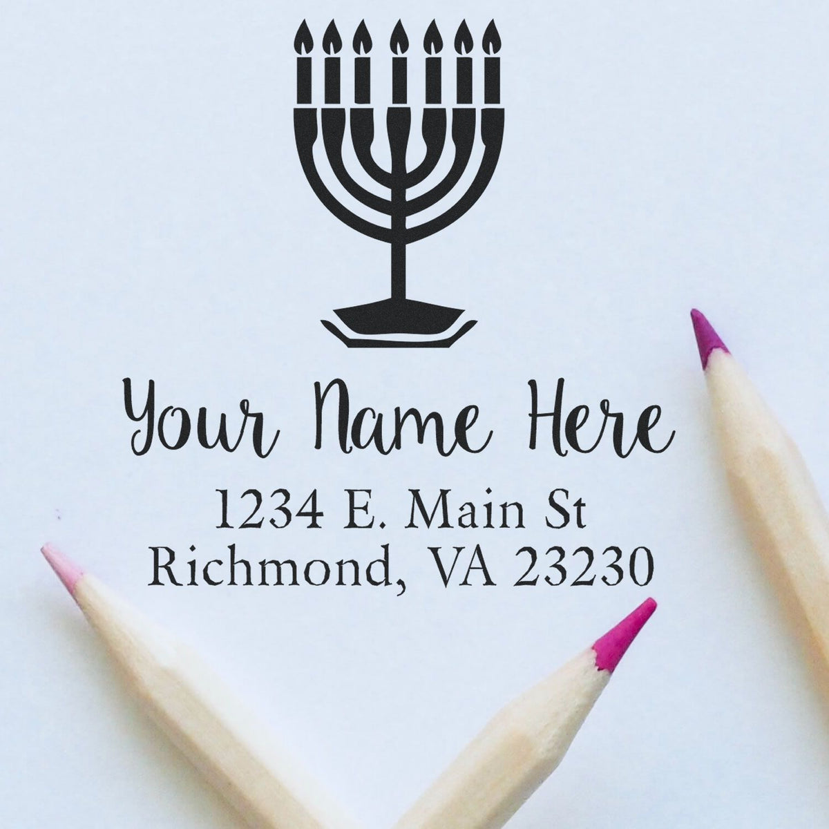 Black menorah stamp and seal with custom name and address on white paper, framed by pink-tipped pencils.