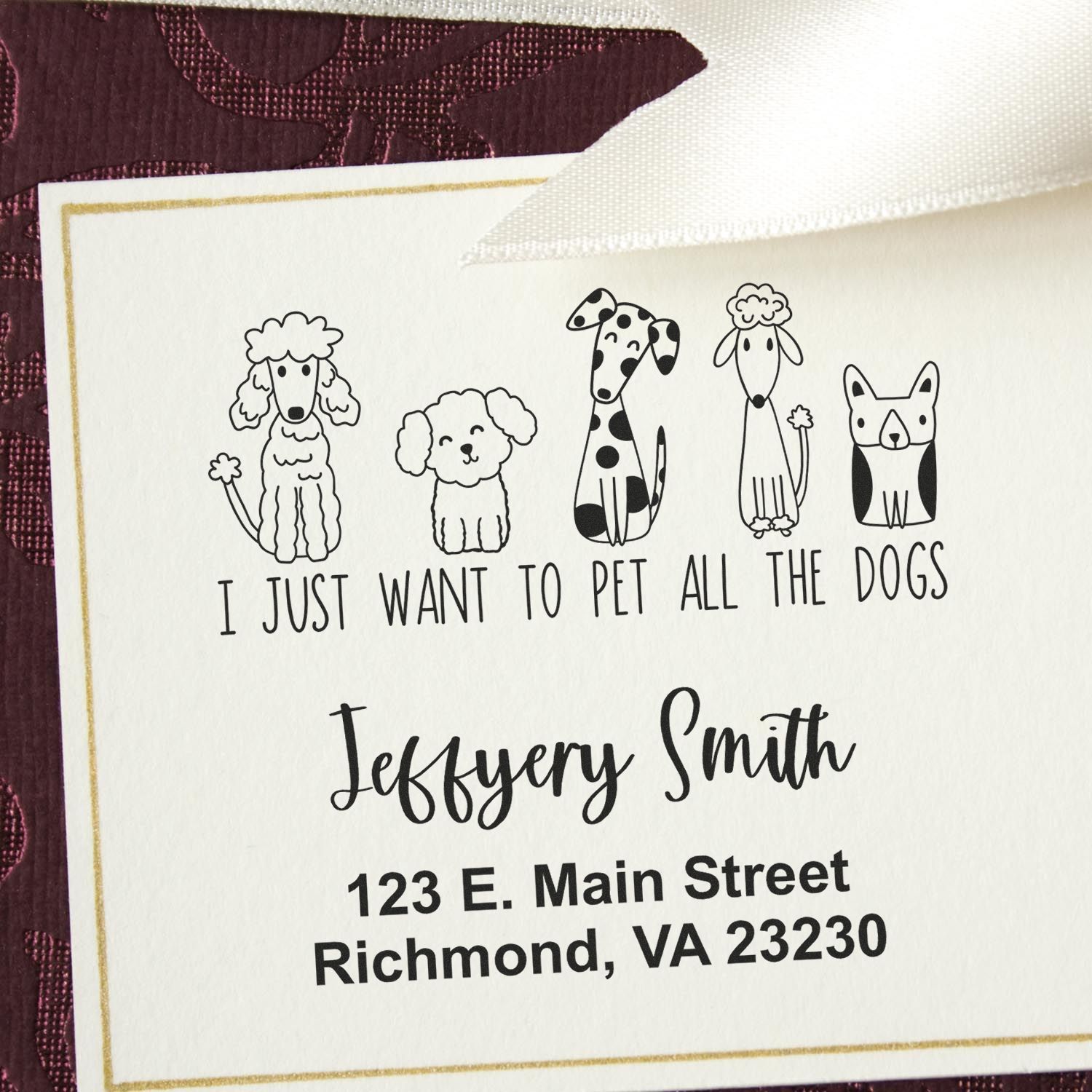 Personalized stamp and seal with dog illustrations, black text, gold border, on a white card over burgundy fabric.