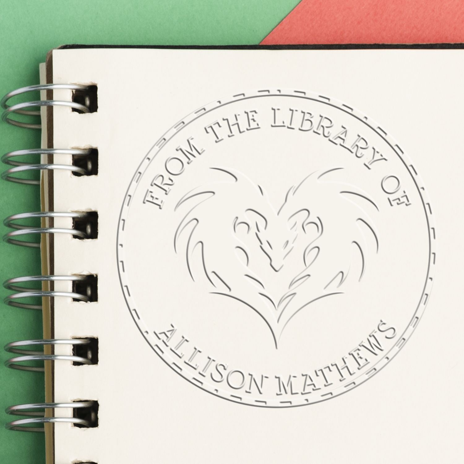 Embossed library seal stamp on cream spiral notebook, with heart-shaped design, silver rings, and green-red background.