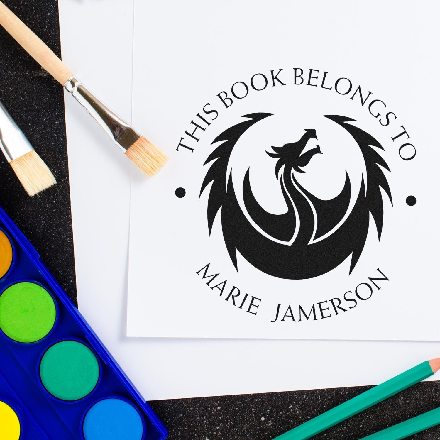 Custom black phoenix book seal stamp on white paper, with paintbrushes, watercolor palette, and teal pencils.
