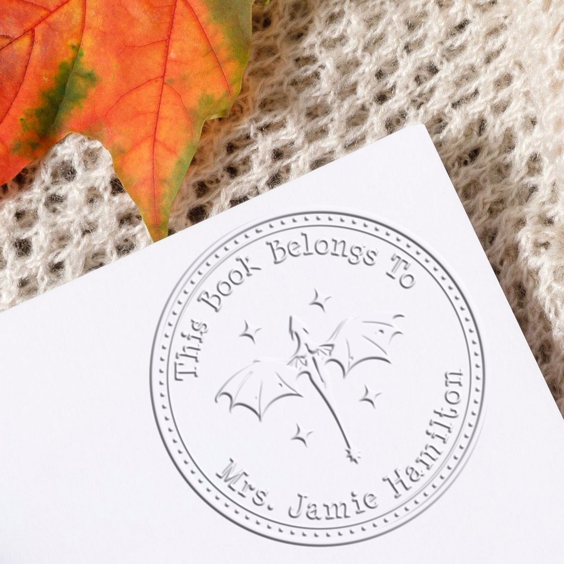 White custom bookplate stamp seal with gray embossed circle, bats, and This Book Belongs To Mrs. Jamie Hamilton, on white paper.