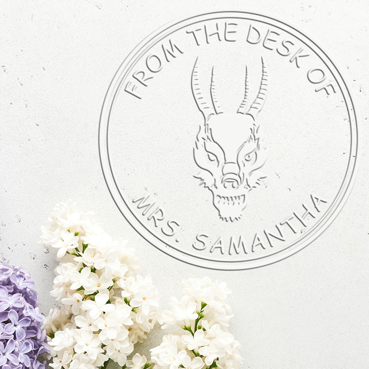 Embossed circular desk stamp seal with goat head, gray text, on white background and white, purple lilac flowers.