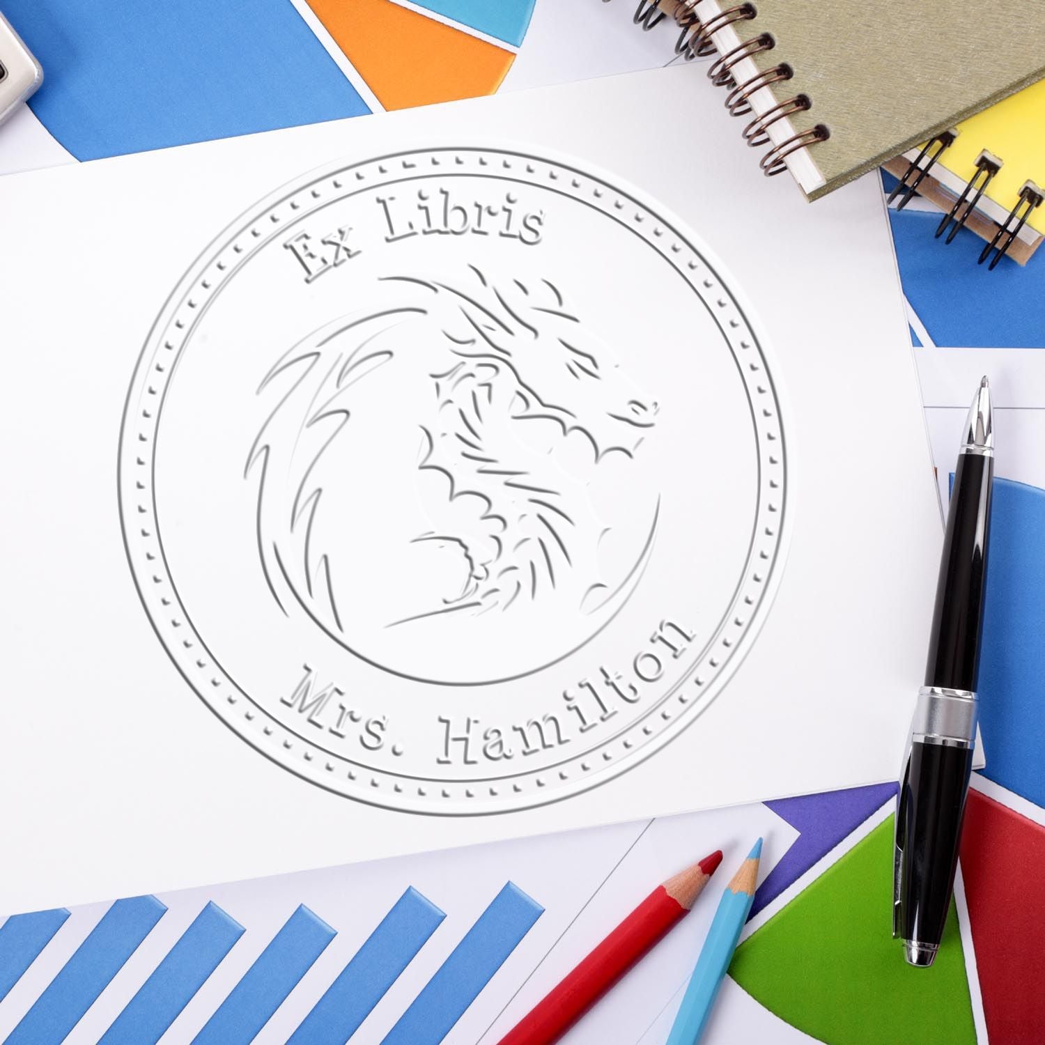 Embossed Ex Libris Mrs. Hamilton library seal on white paper, surrounded by colorful stationery, pens, pencils, and notebooks.