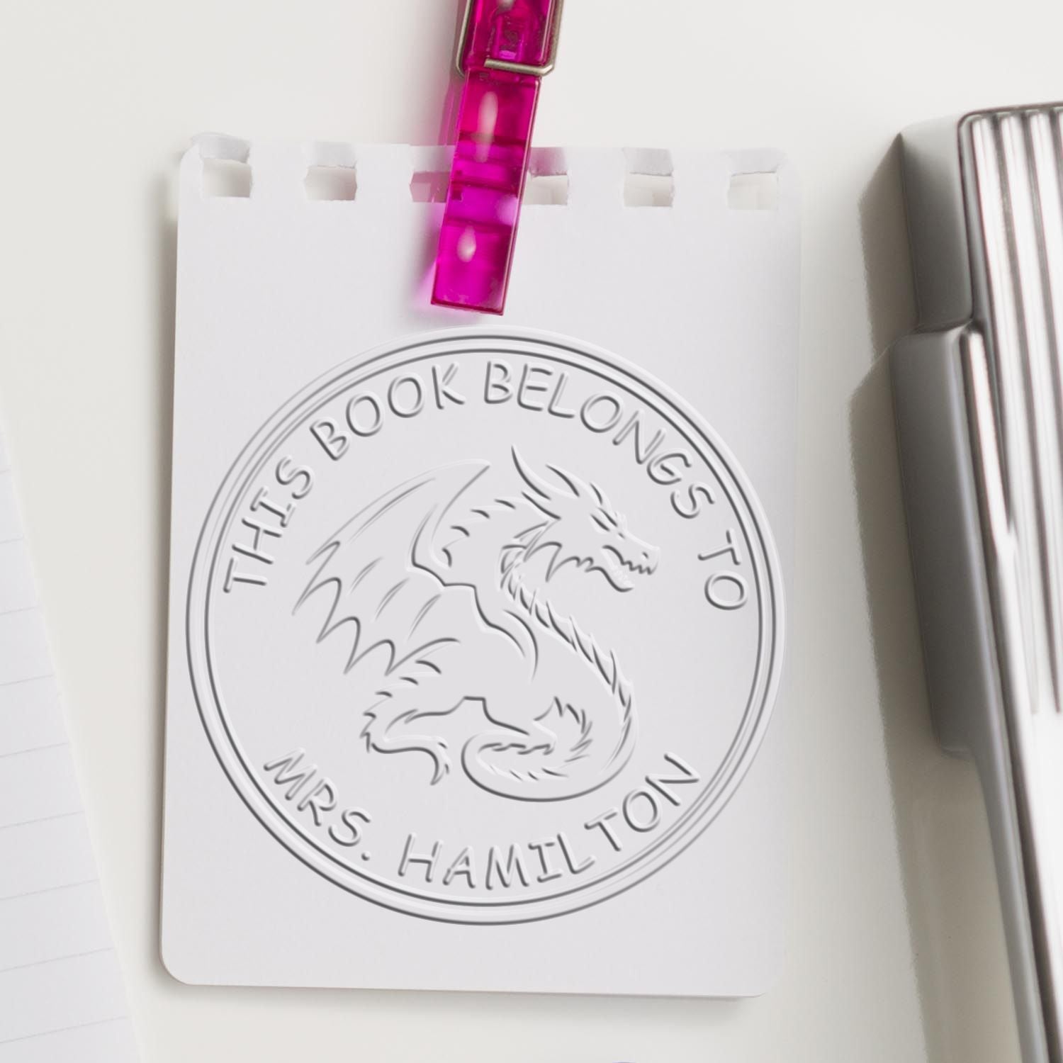 White bookplate stamp and seal card with embossed dragon seal for Mrs. Hamilton, clipped with pink clip beside silver stamp.