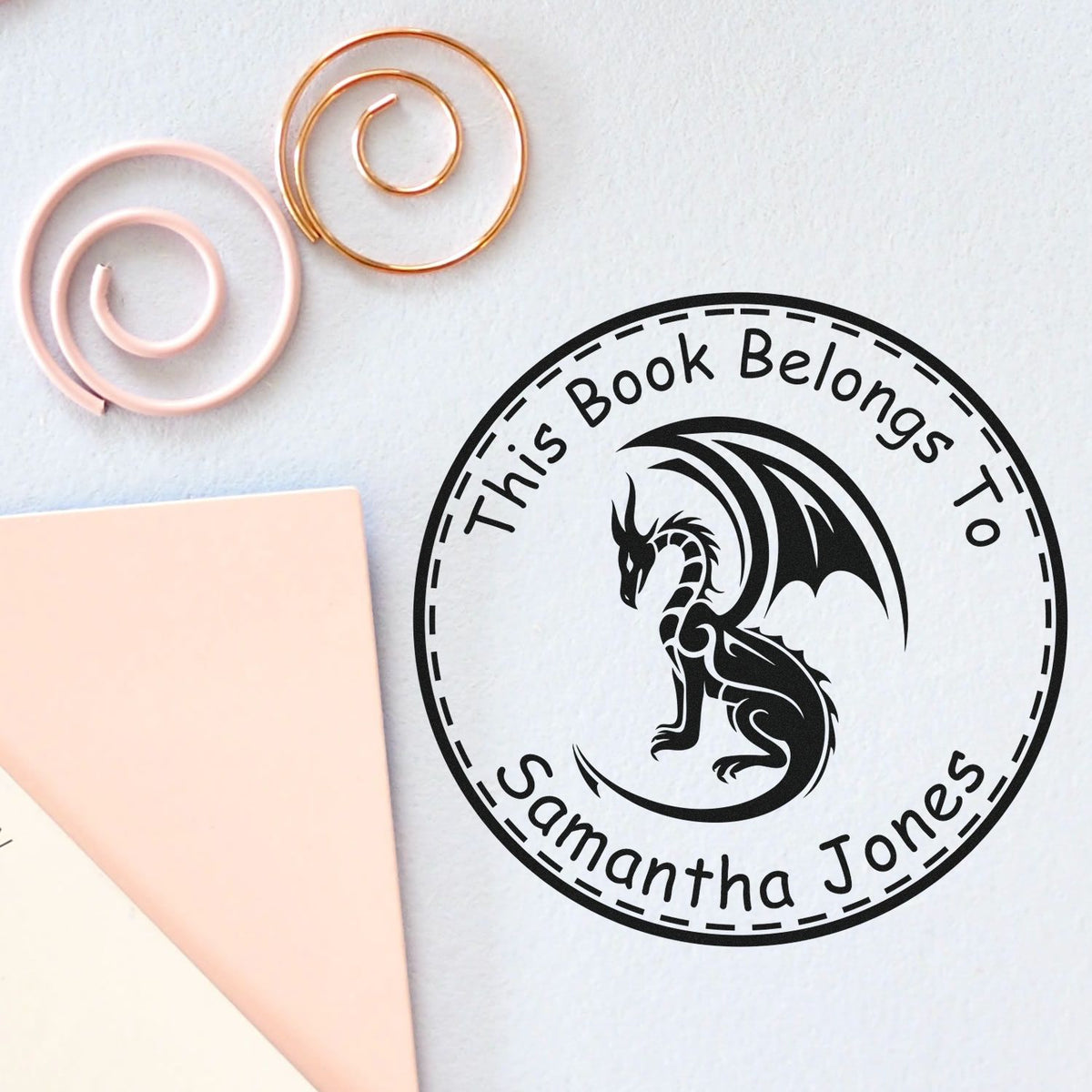 Custom black book-ownership seal stamp with dragon, surrounded by pastel pink and gold spiral clips on white background
