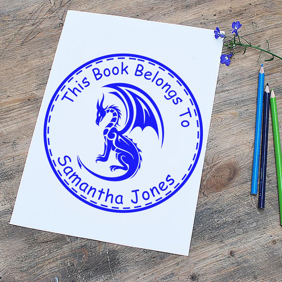 Blue custom book stamp seal with dragon and This Book Belongs To Samantha Jones on white paper beside pencils and flowers.