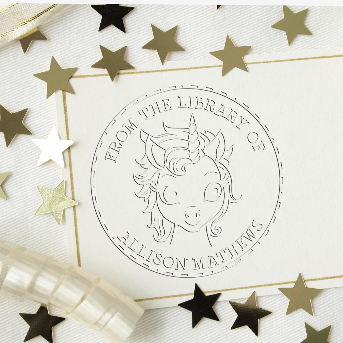 White embossed unicorn library stamp seal with gold trim, surrounded by black and gold star confetti.