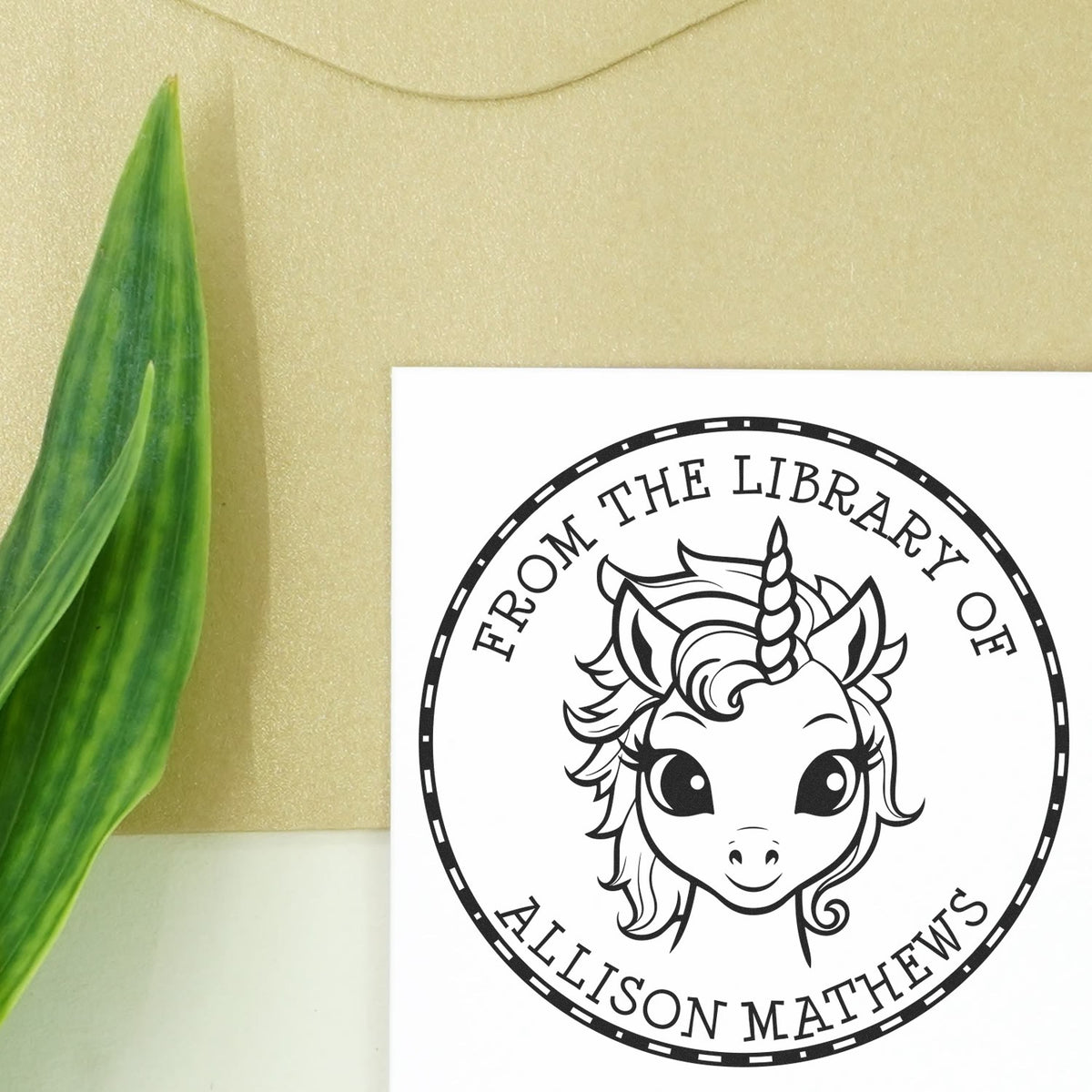 Black custom library stamp and seal featuring a unicorn head in a circular border on a white card, beside a green leaf.