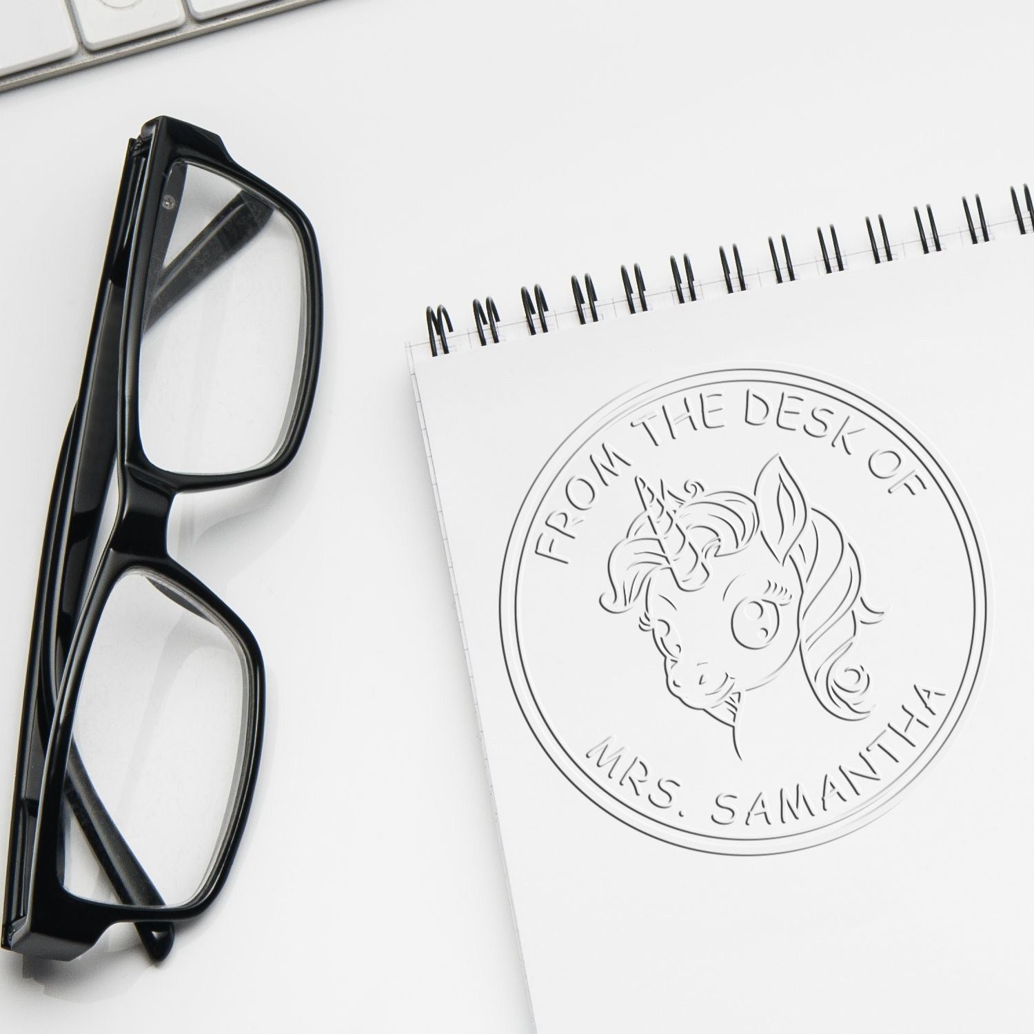Black desk stamp and embossed seal on white notebook beside black eyeglasses, featuring a unicorn Mrs. Samantha design.