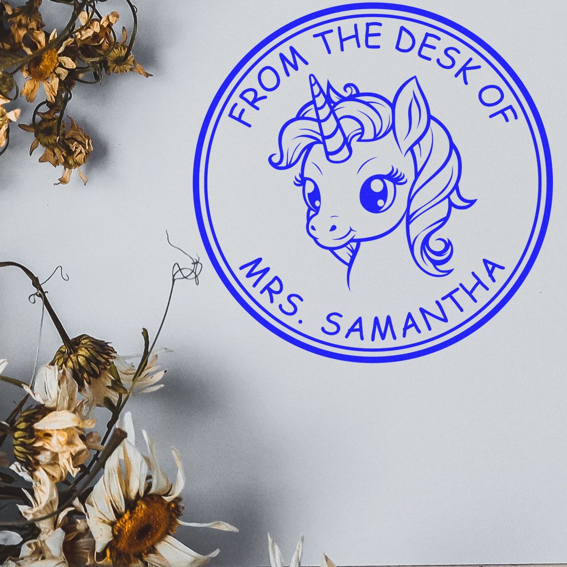 Blue circular unicorn stamp seal reading From the Desk of Mrs. Samantha on a light background with dried flowers.