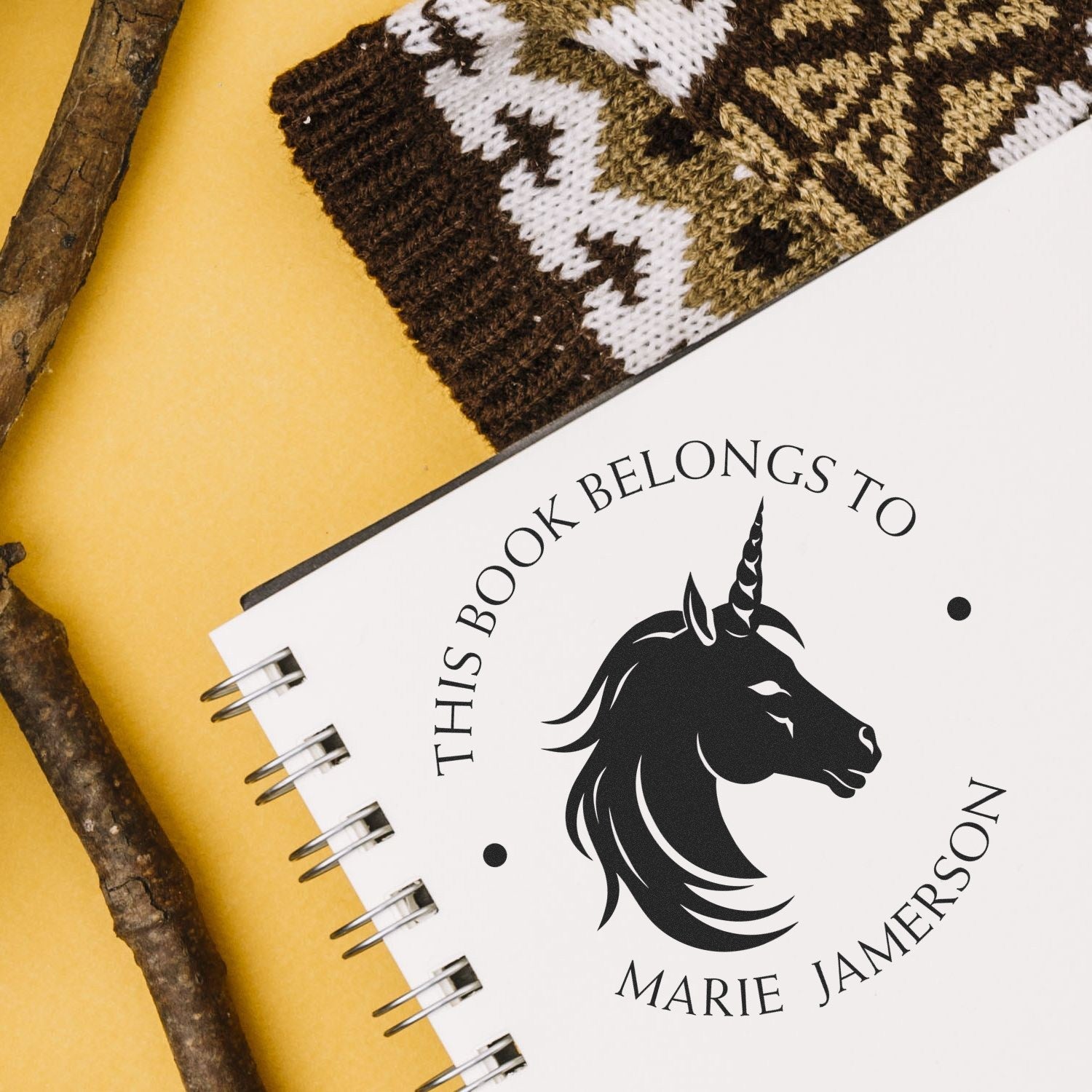 Personalized black unicorn book stamp and seal on a white spiral notebook, with mustard yellow background and brown knit fabric.