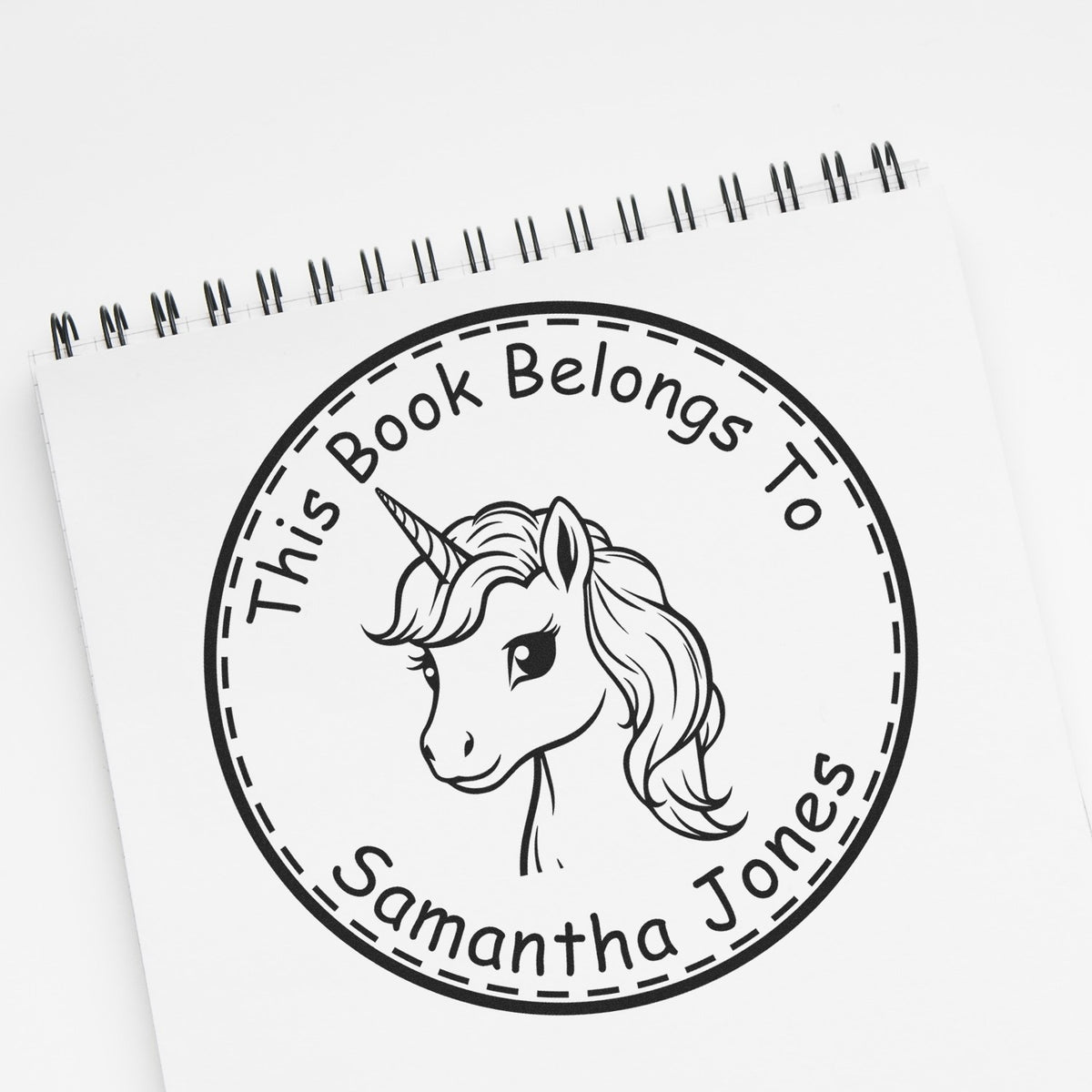 Black custom unicorn book seal stamp with This Book Belongs To Samantha Jones on a white spiral notepad.