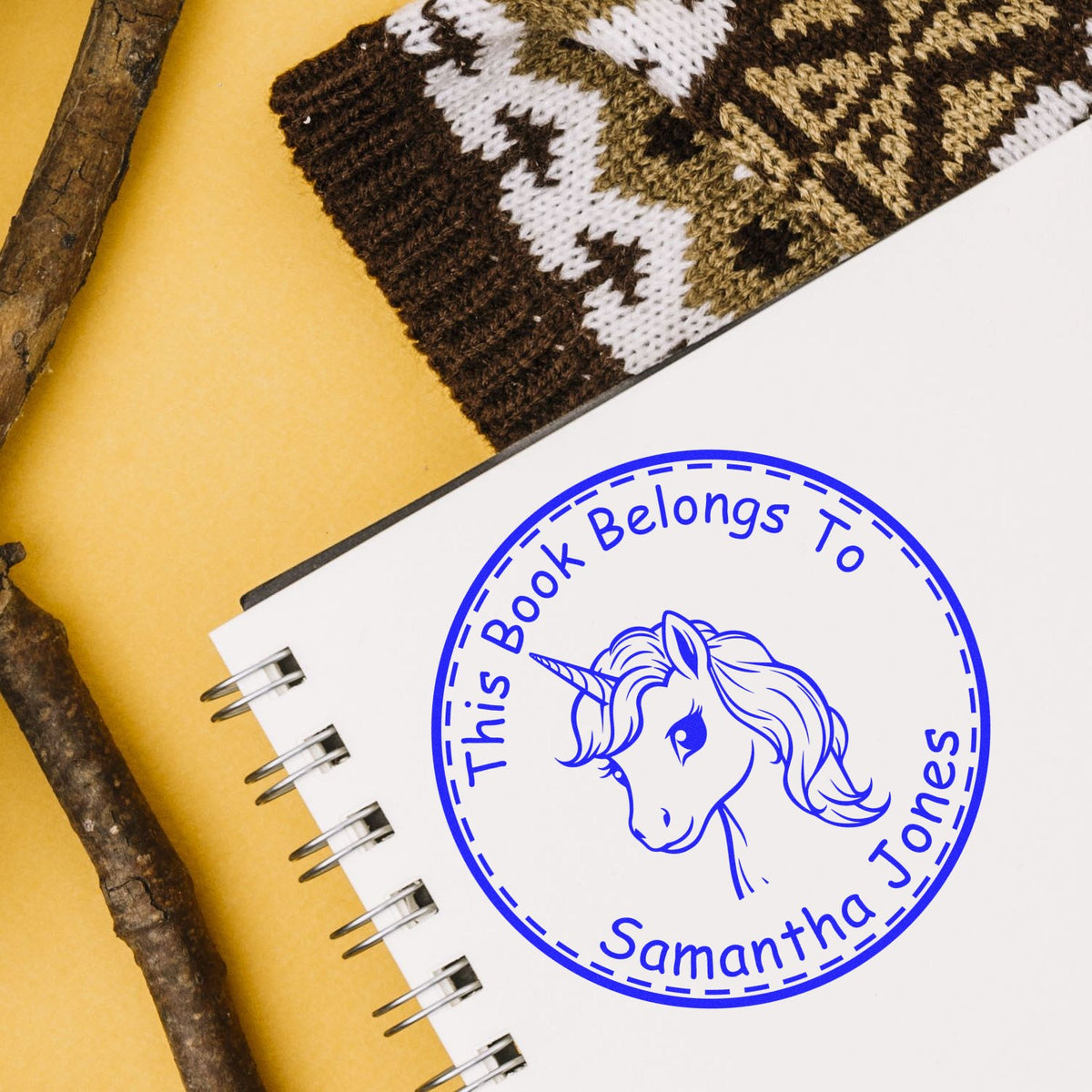 Blue personalized unicorn bookplate stamp seal on white notebook, labeled This Book Belongs to Samantha Jones, on yellow with brown knit accents.