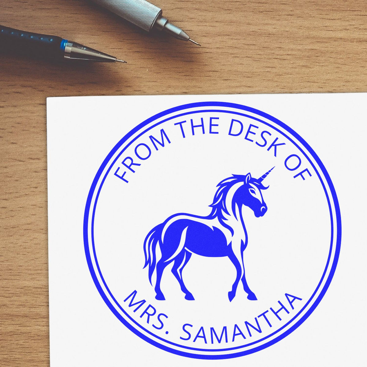 Blue custom desk seal stamp with unicorn and From the Desk of Mrs. Samantha, on white paper beside two pens.