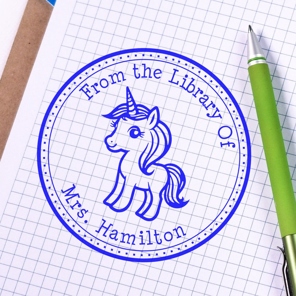 Blue circular library stamp seal with unicorn and Mrs. Hamilton text on graph paper, beside a green pen.