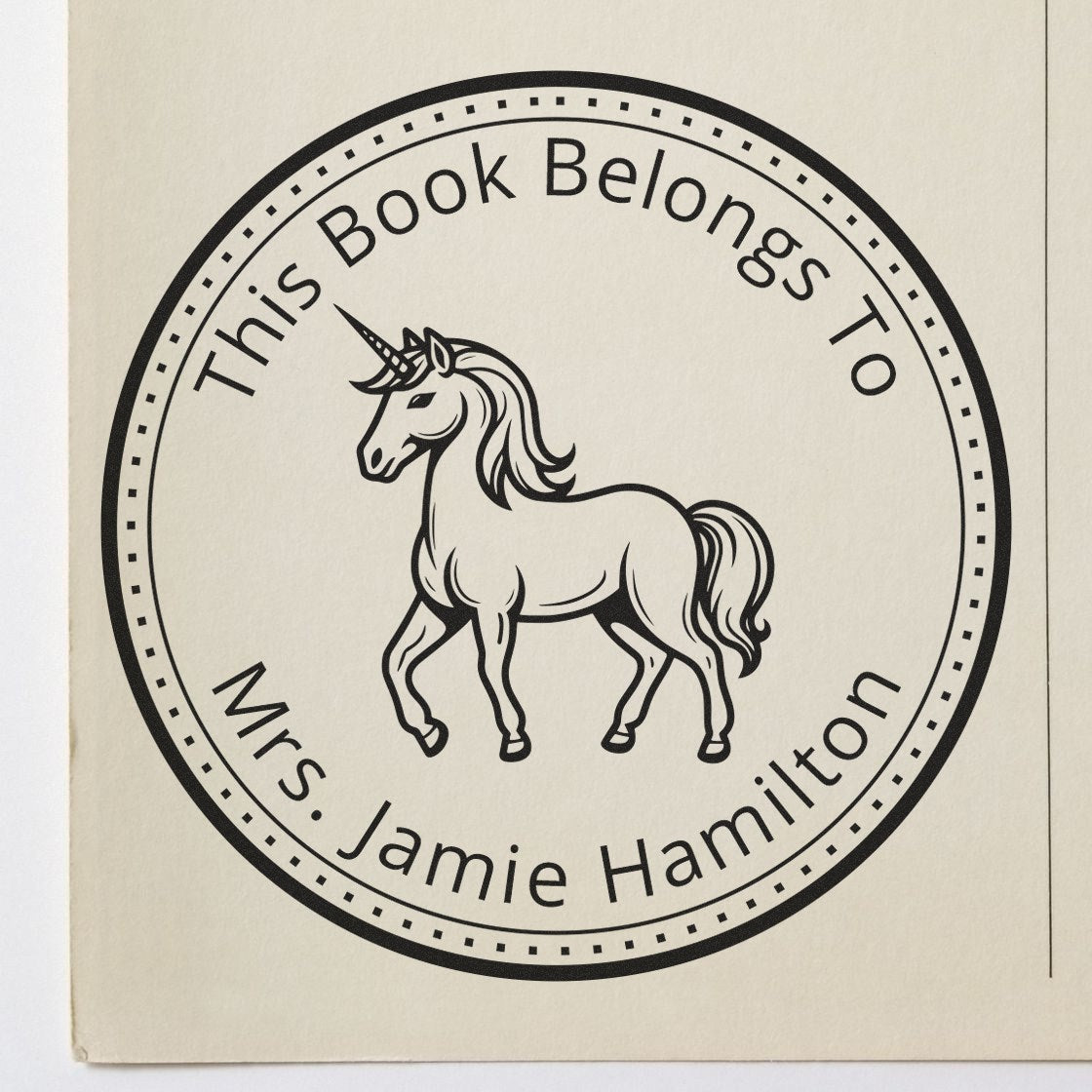 Black circular custom book stamp seal with unicorn and text This Book Belongs To Mrs. Jamie Hamilton on cream paper.