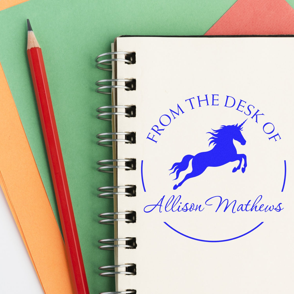 Custom blue unicorn desk seal stamp reading Allison Mathews on a white spiral notebook, with red pencil and green-orange backdrop
