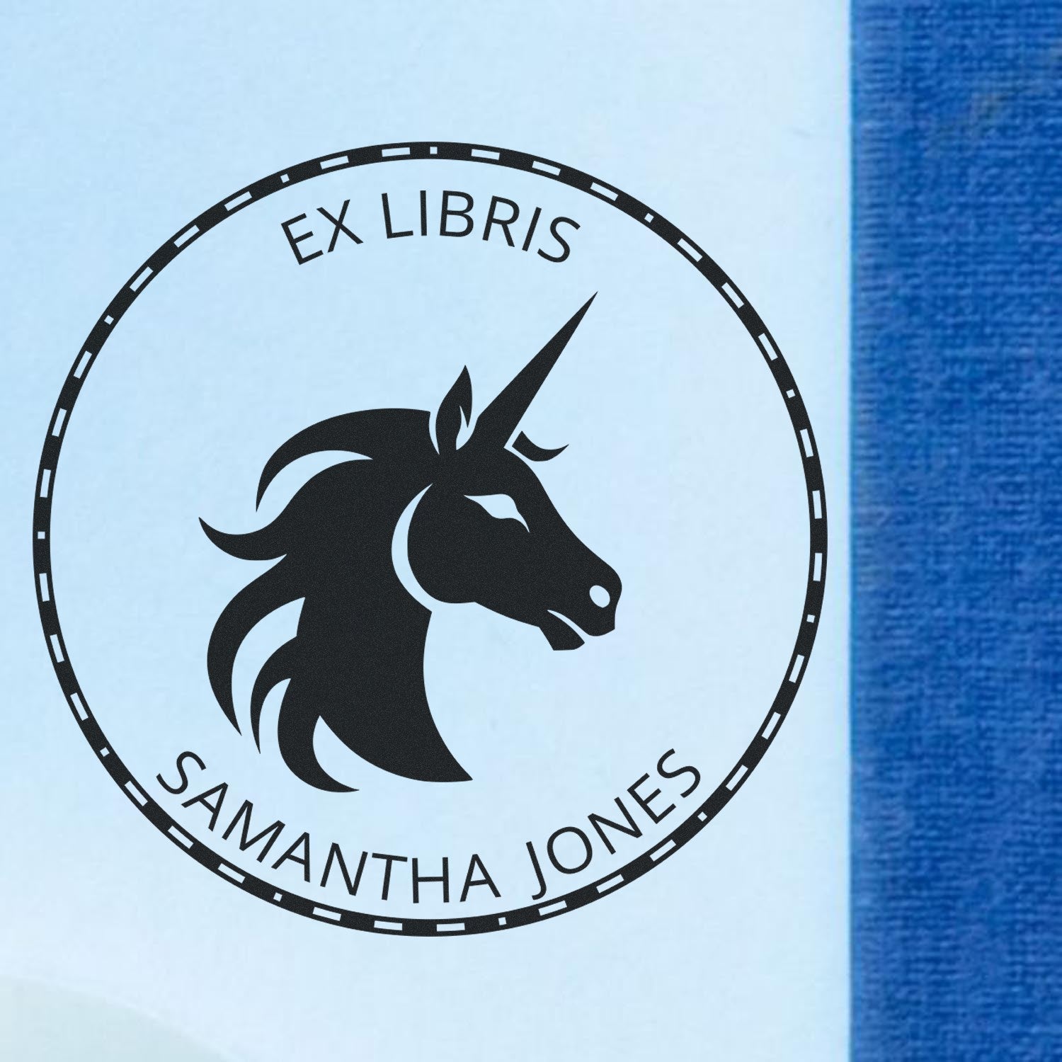 Black circular ex libris stamp seal with unicorn head and Samantha Jones text on white paper beside blue fabric.
