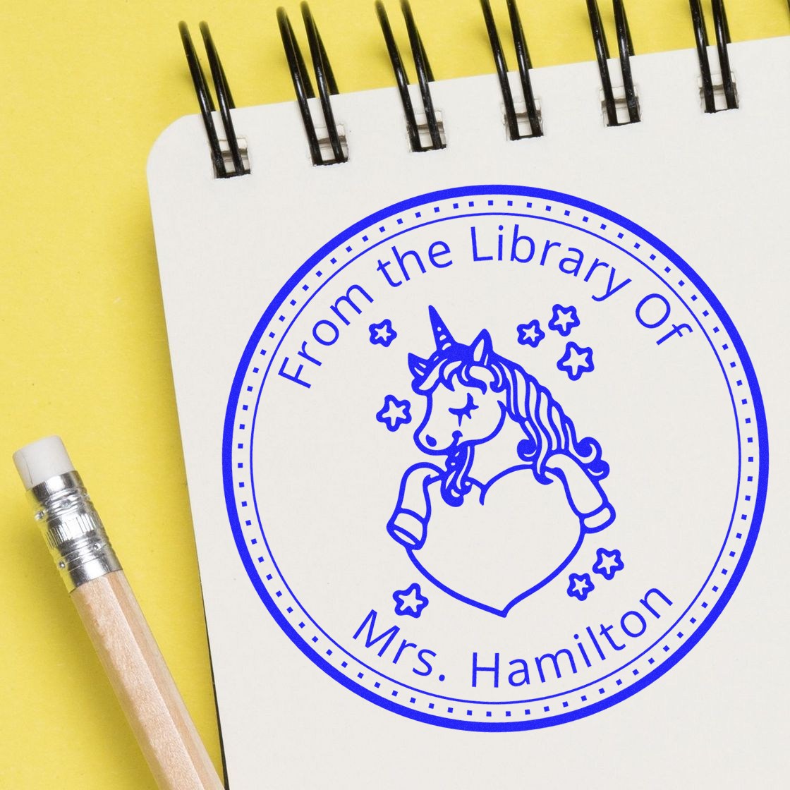 Blue custom library stamp seal with unicorn and stars on a white notebook, beside a pencil on a yellow background.