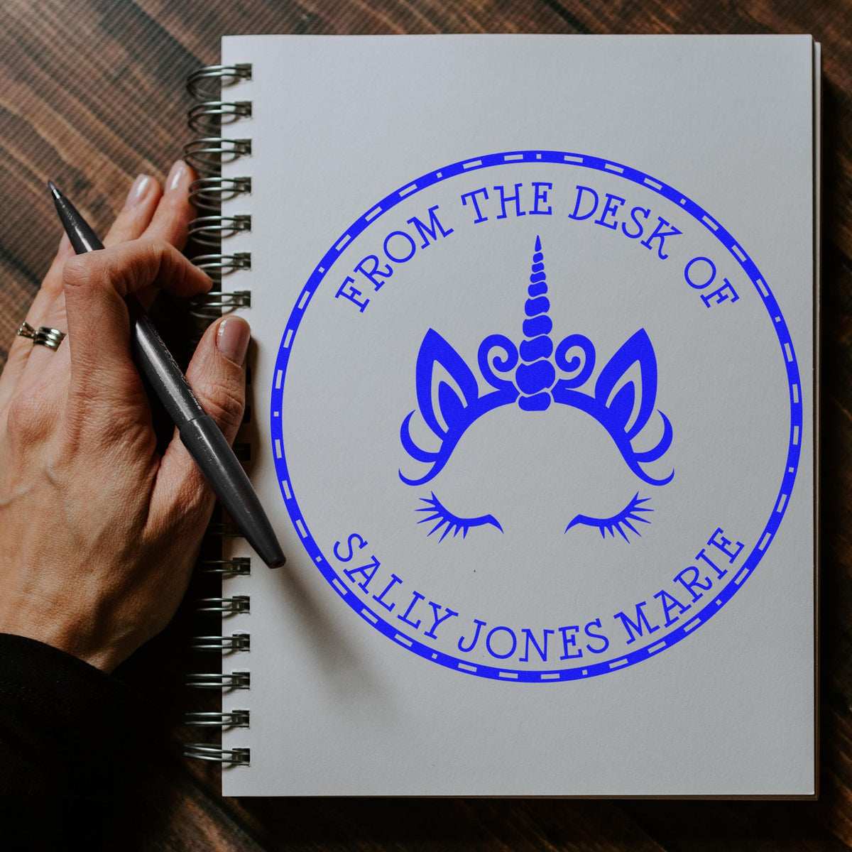 Hand holding pen beside a spiral notebook with a blue unicorn custom stamp seal reading Sally Jones Marie.