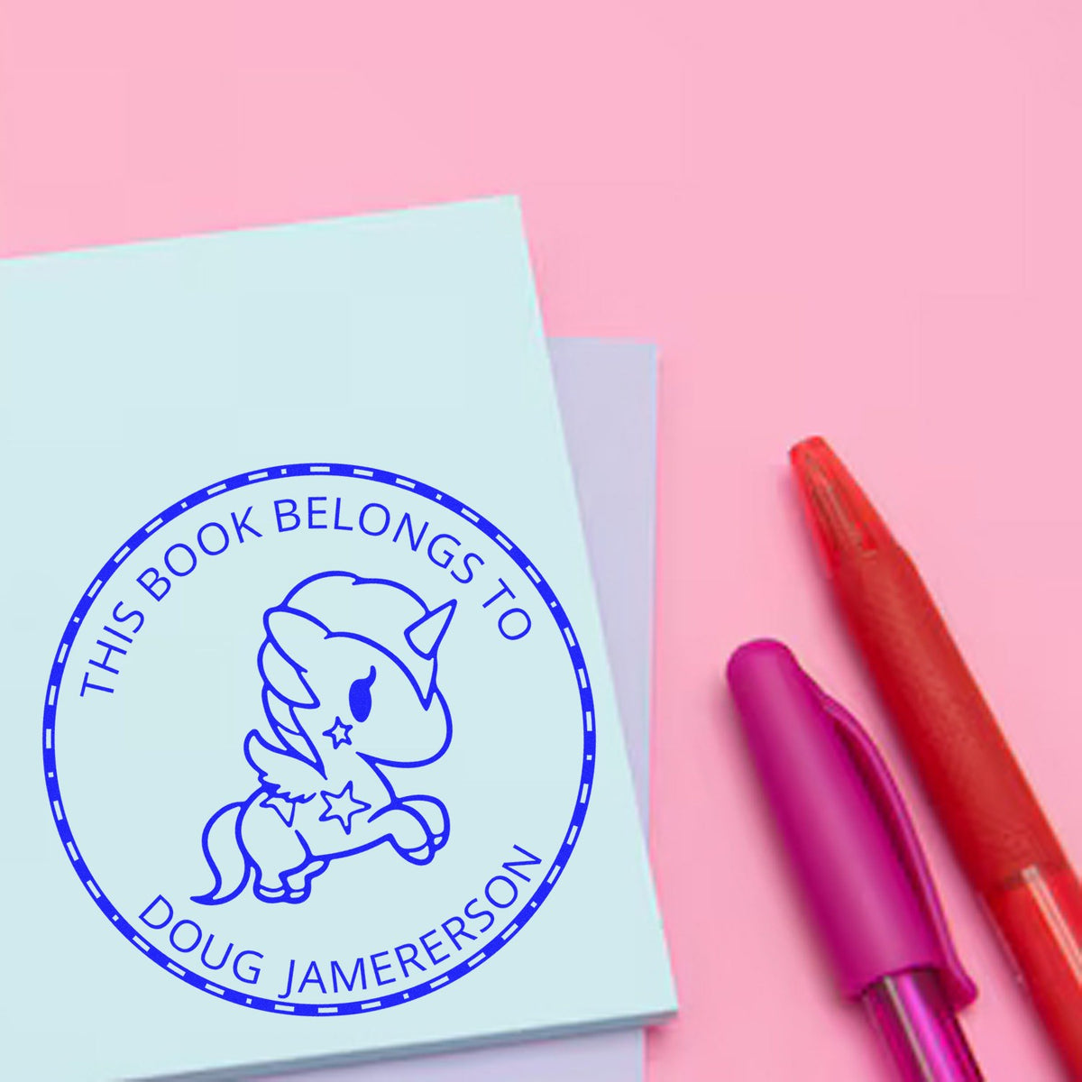 Blue unicorn book-belongs-to stamp seal on pastel pink paper, beside red and purple pens.