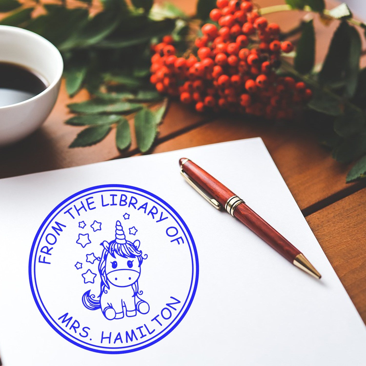 Blue circular library stamp seal with unicorn and From the Library of Mrs. Hamilton on white paper, beside a burgundy pen.