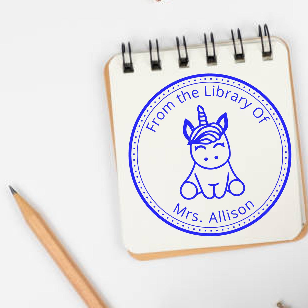Blue circular unicorn library stamp seal on a cream spiral notepad, with Mrs. Allison and a pencil.