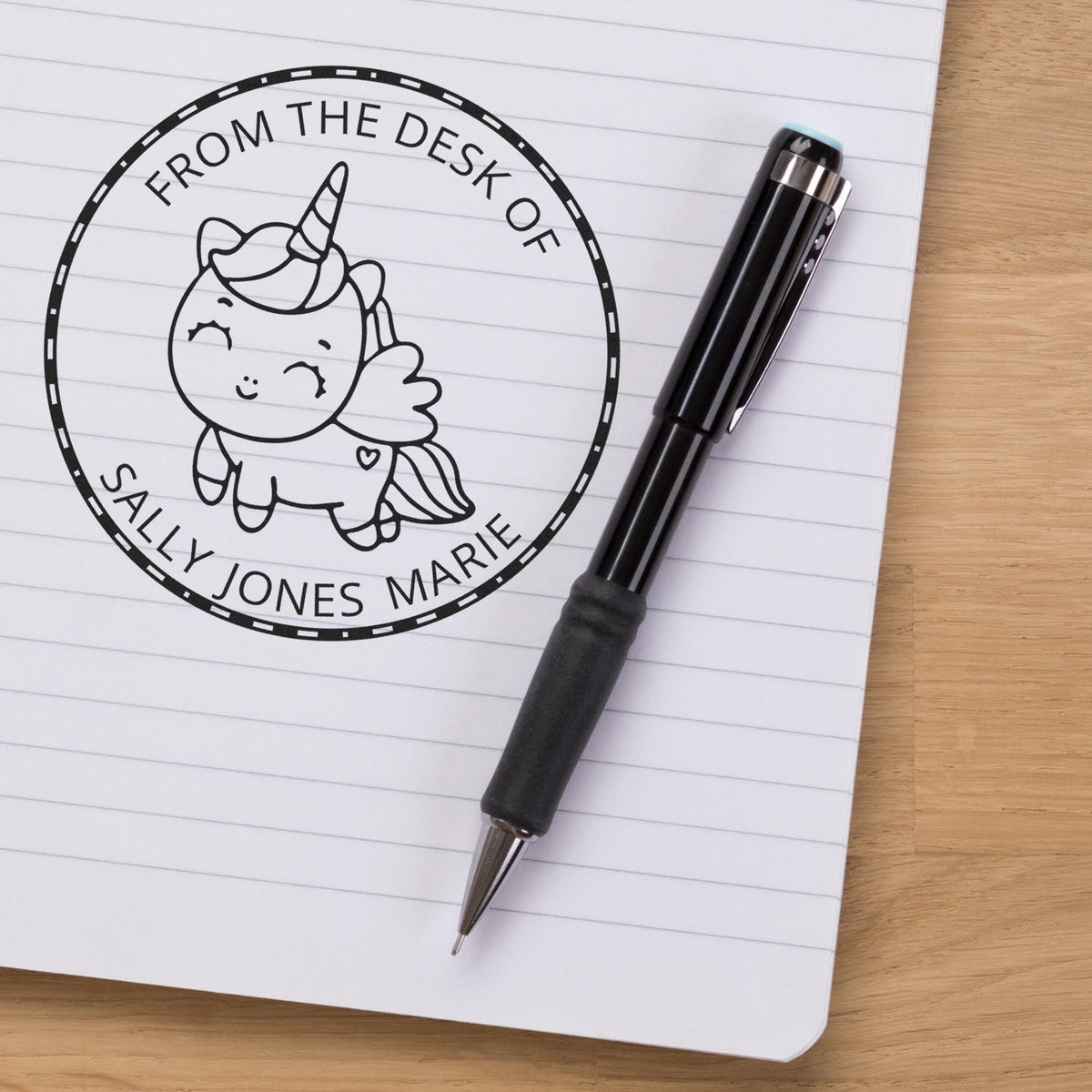 Custom black unicorn desk seal stamp reading From the Desk of Sally Jones Marie beside a black pen on a lined notebook.