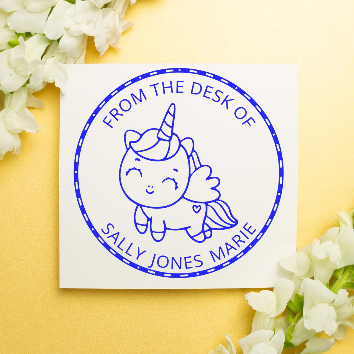 Blue unicorn desk stamp seal with Sally Jones Marie text on white square, set on soft yellow background with white flowers.