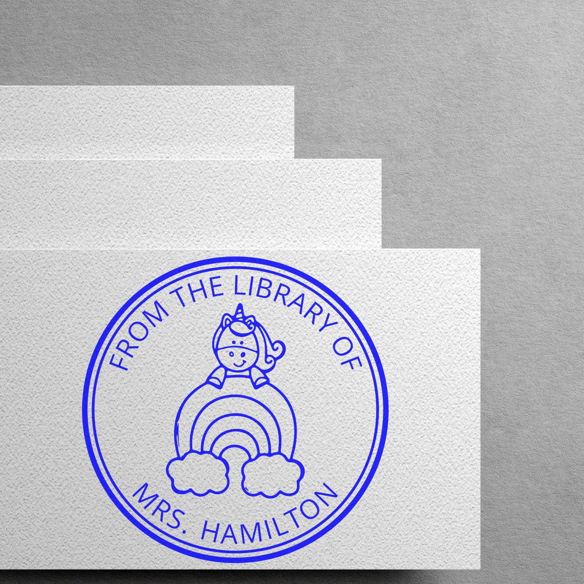 Blue custom library stamp seal with unicorn and rainbow, reading From the Library of Mrs. Hamilton on textured white paper.