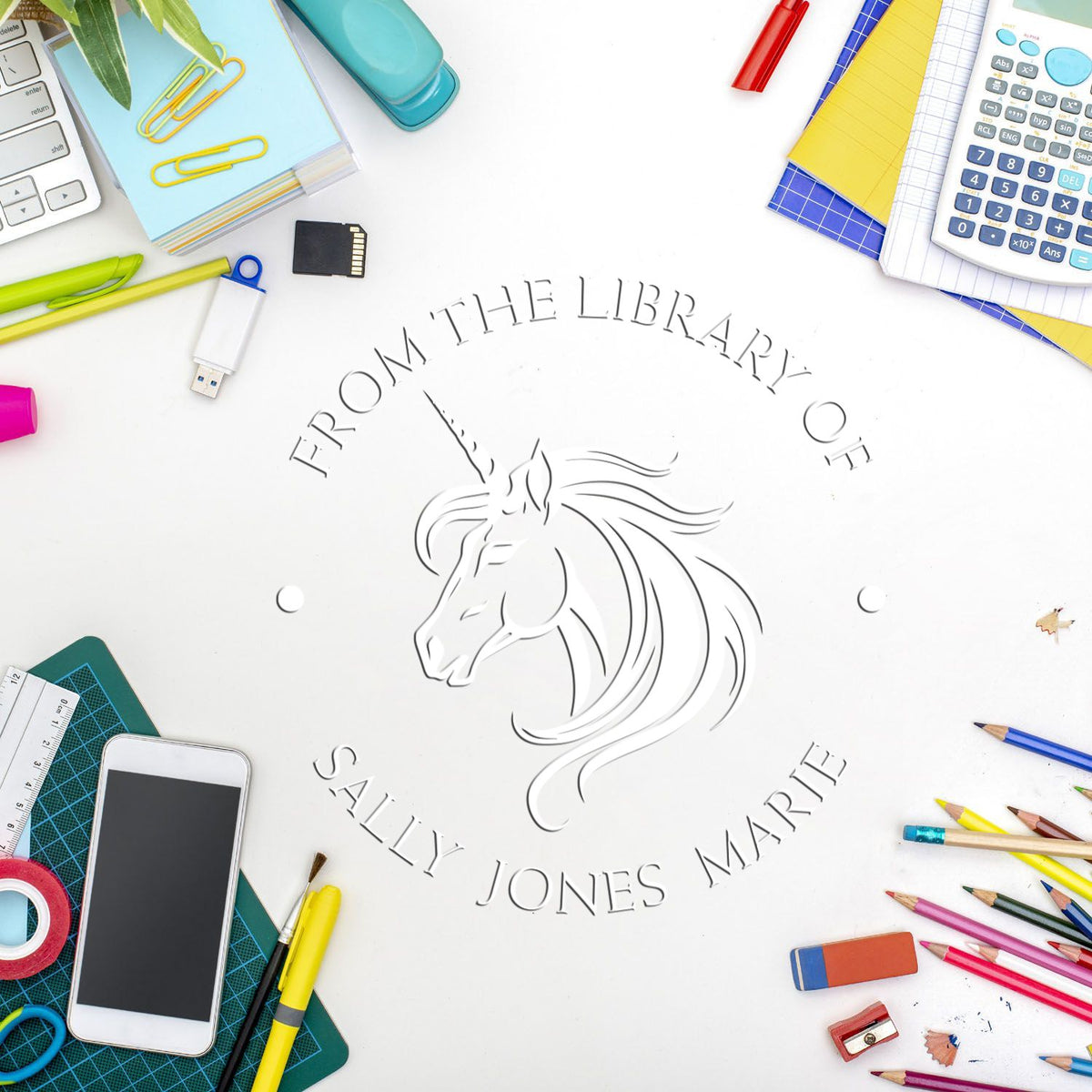 Custom unicorn library stamp and embossing seal on white desk, surrounded by colorful stationery, pencils, calculator, and phone.