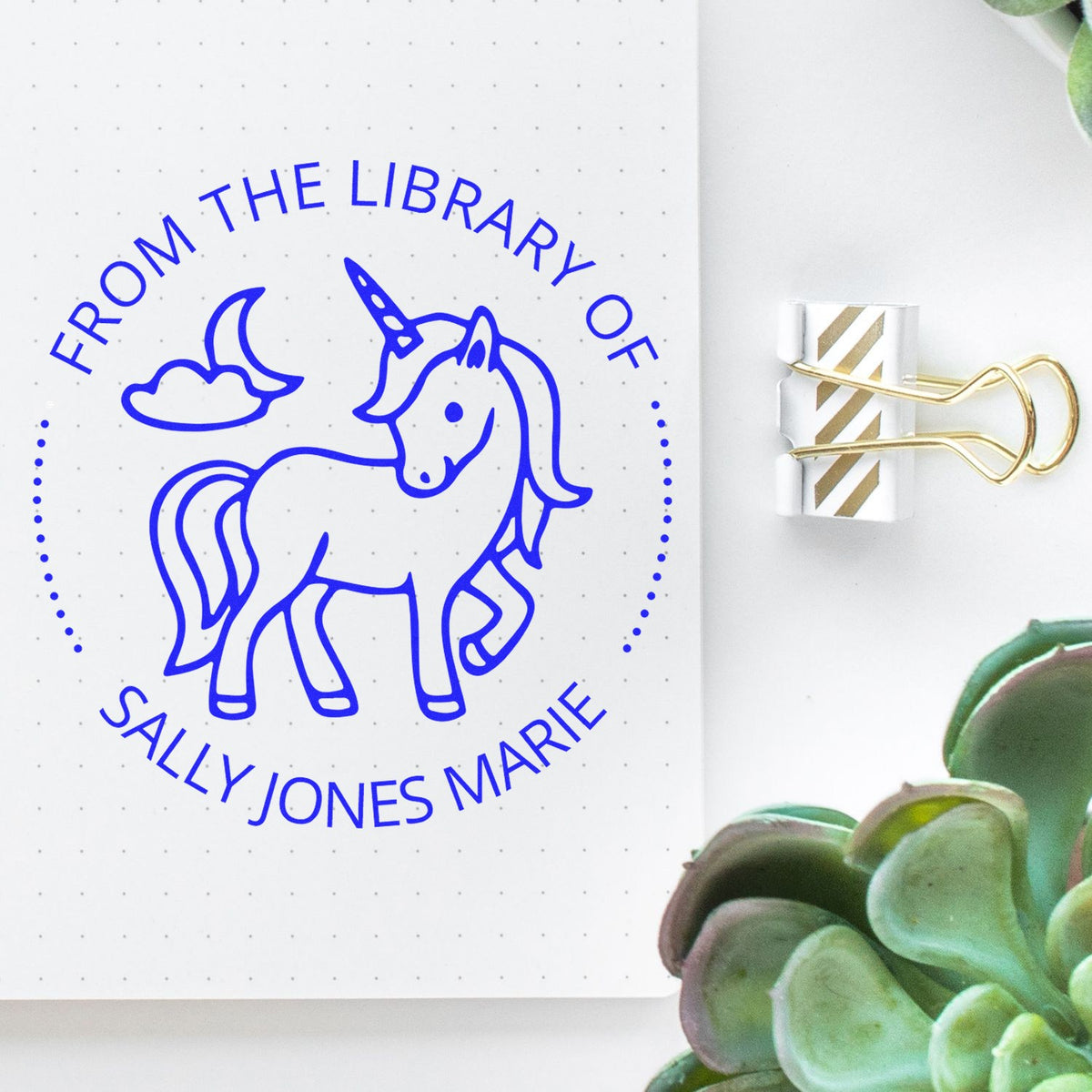 Blue unicorn library stamp on white dotted notebook, with a gold-striped binder clip seal and succulents.