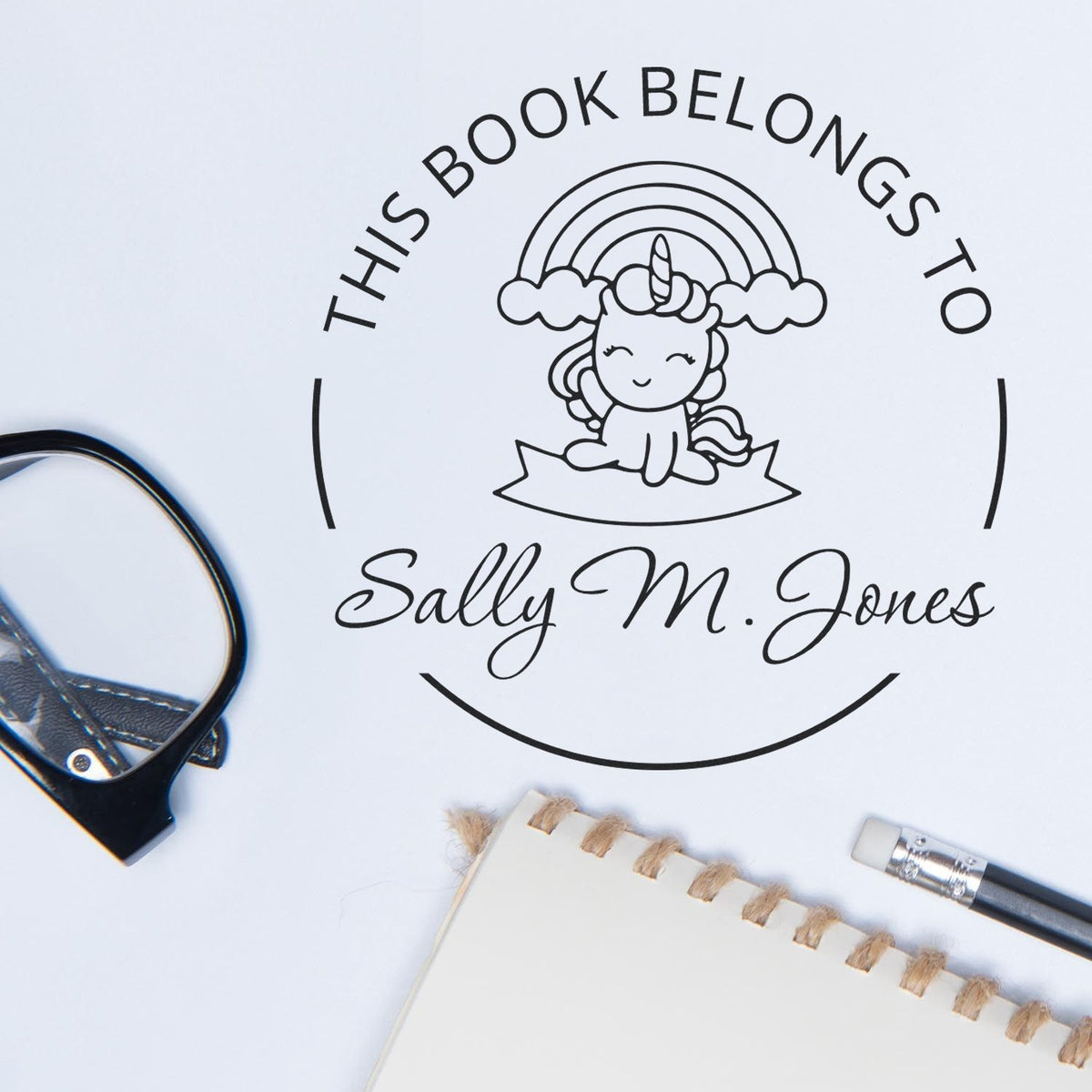 Black book-belongs-to stamp with unicorn seal and Sally M. Jones text on white, beside notebook, pen, and clip.