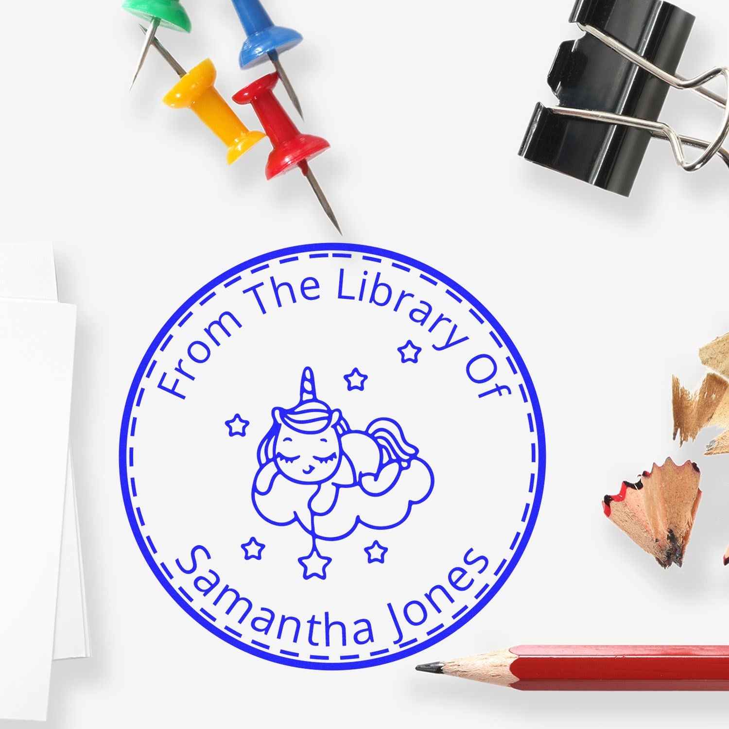 Blue custom library seal stamp with unicorn, stars, and Samantha Jones, surrounded by colorful push pins, binder clip, and pencils.