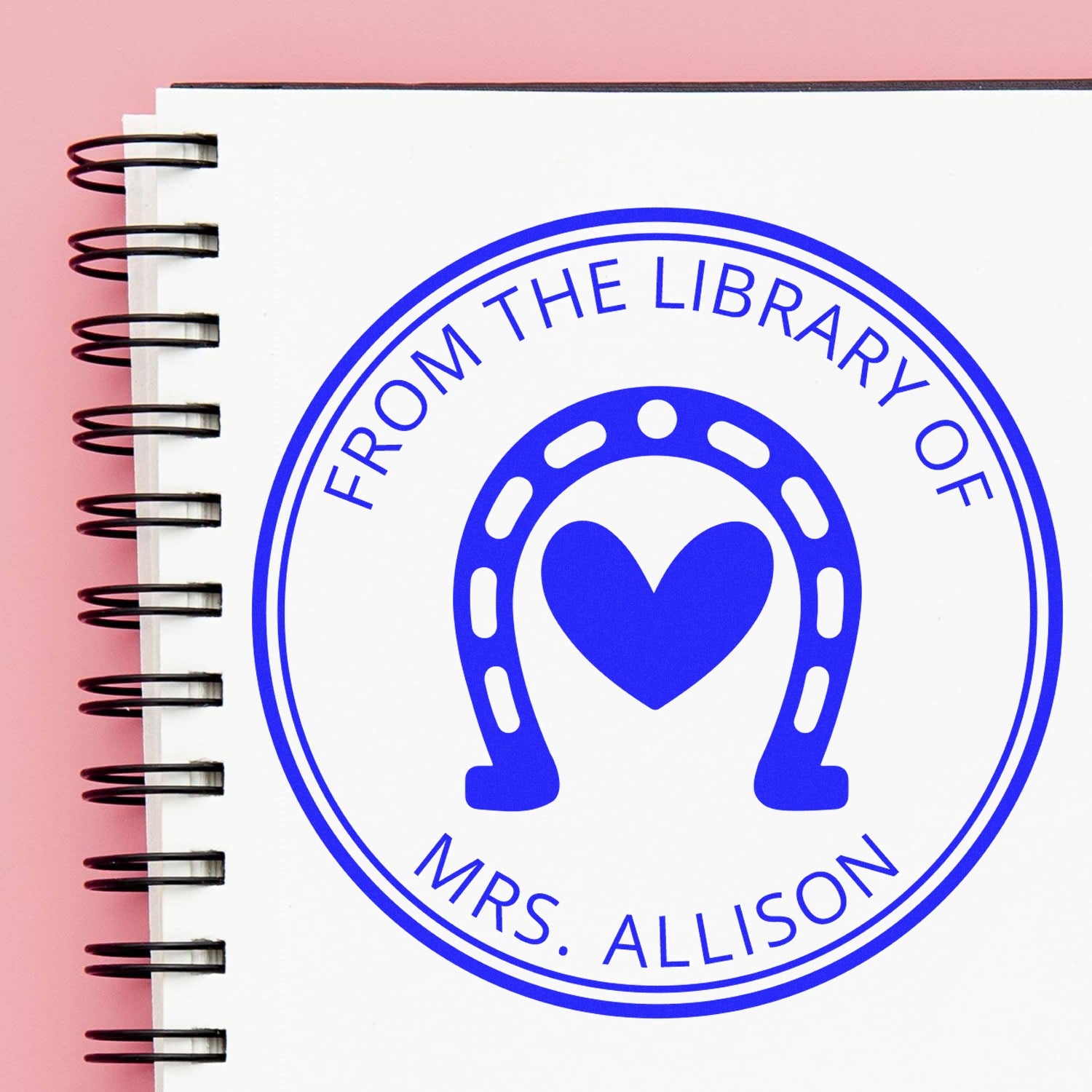 Blue custom library seal stamp on a white spiral notebook, with heart and horseshoe icon, against a pink background.