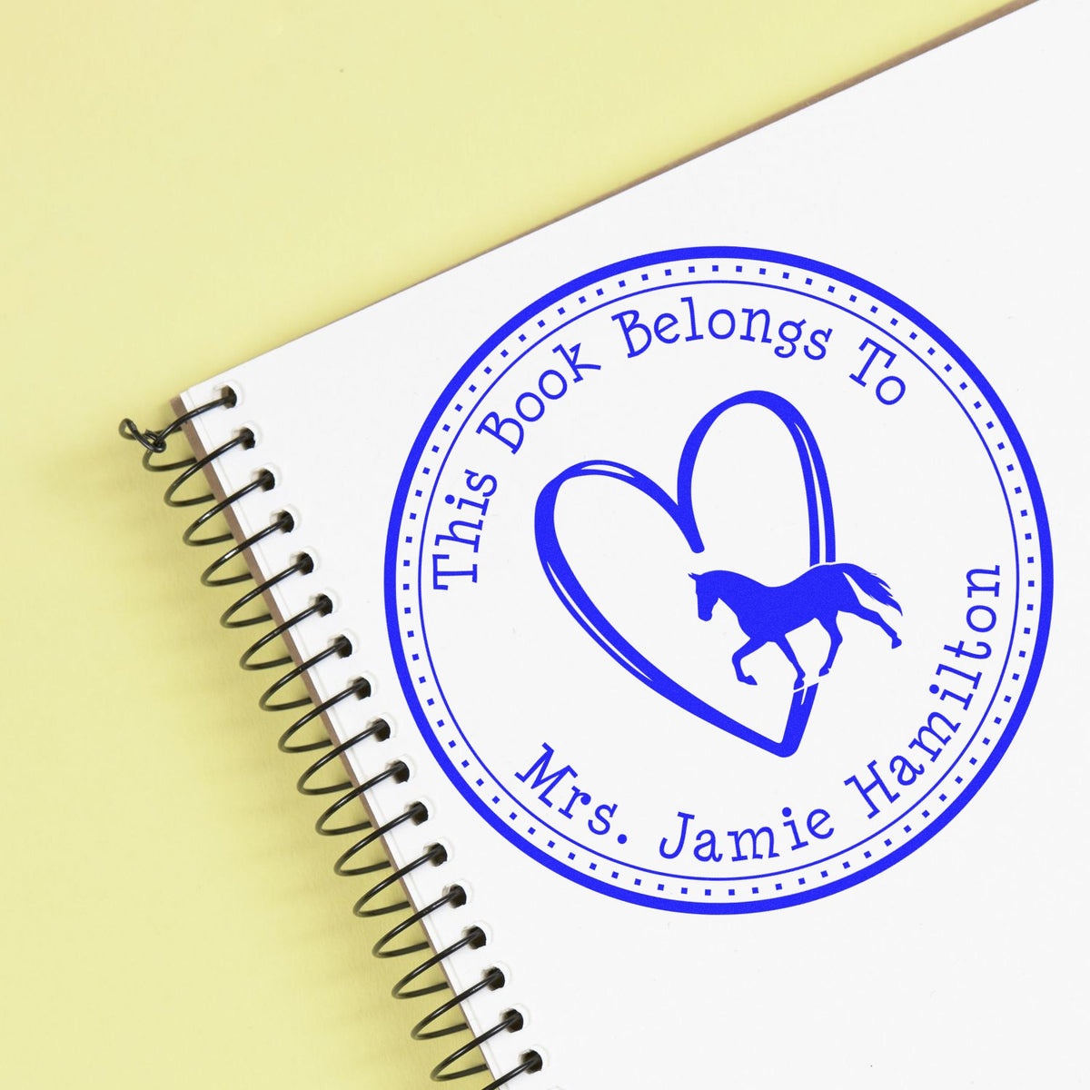 Blue circular book ownership stamp seal with horse in a heart, on a white spiral notebook against a pale yellow background.