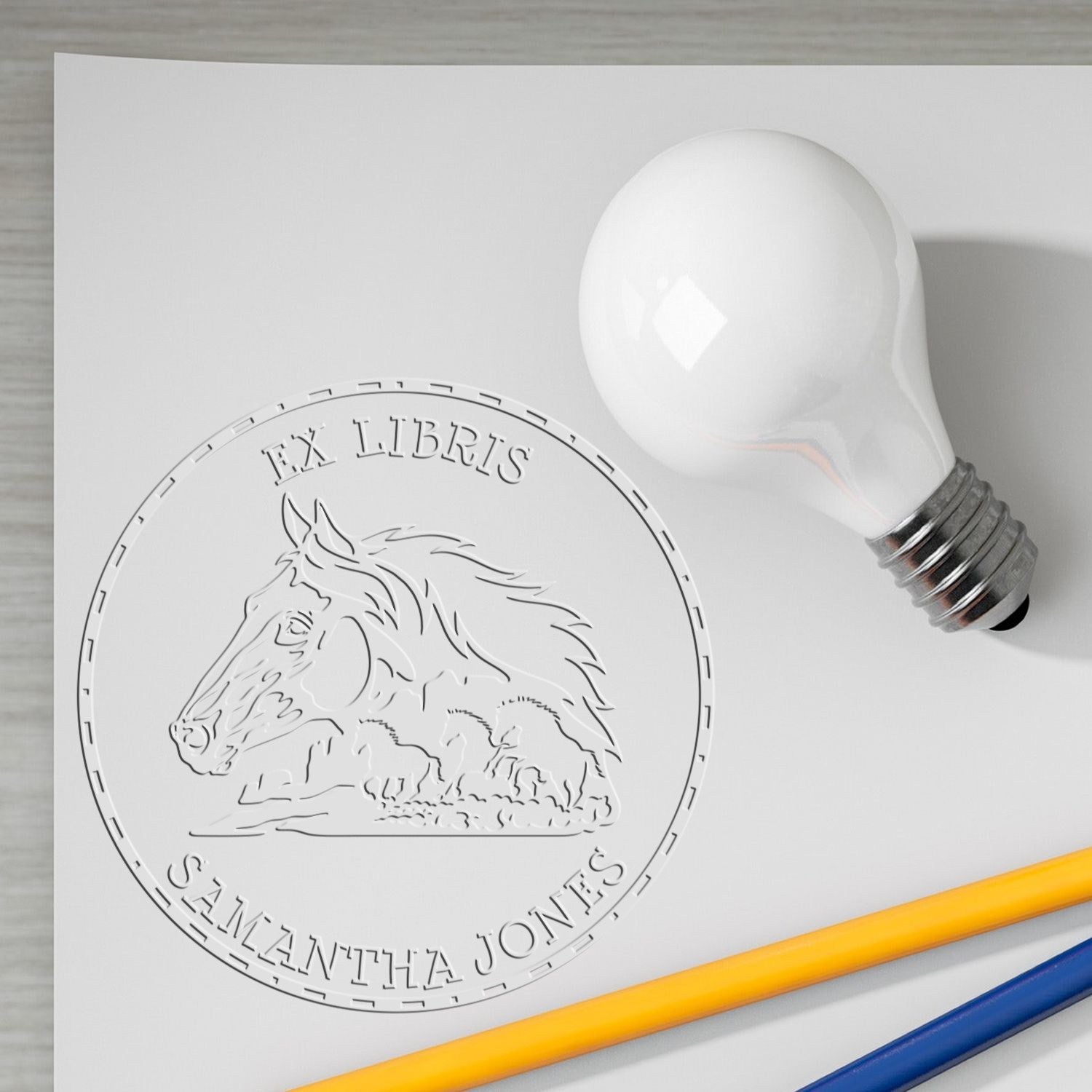 White light bulb stamp and circular seal with horse emblem and engraved text on paper, plus yellow and blue pencils.
