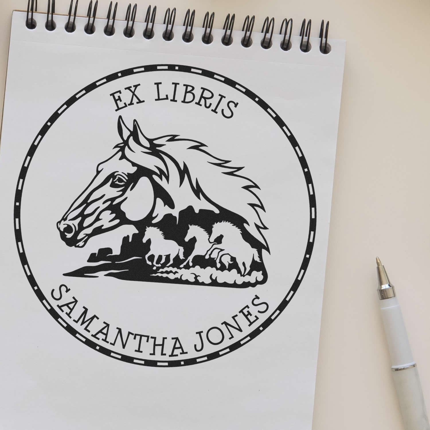 Black ex libris stamp seal with horse head and Samantha Jones on a white spiral notebook, beside a white pen.