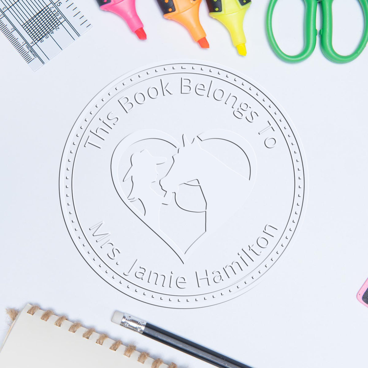 White custom book ownership seal stamp with heart and profiles, surrounded by colorful highlighters, ruler, scissors and pencil.