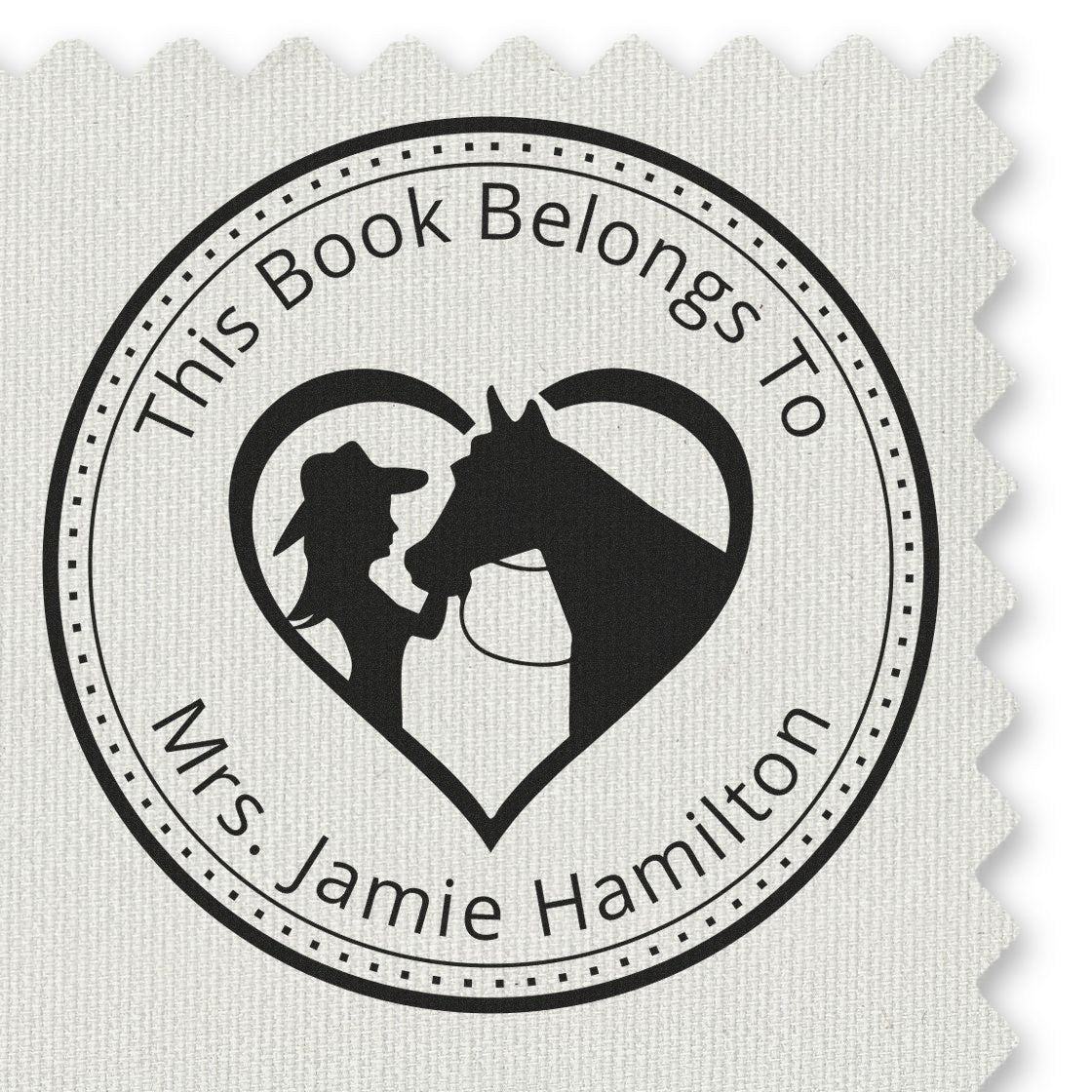 Round black stamp seal with heart and horse silhouette, reading This Book Belongs To Mrs. Jamie Hamilton, on white textured fabric.