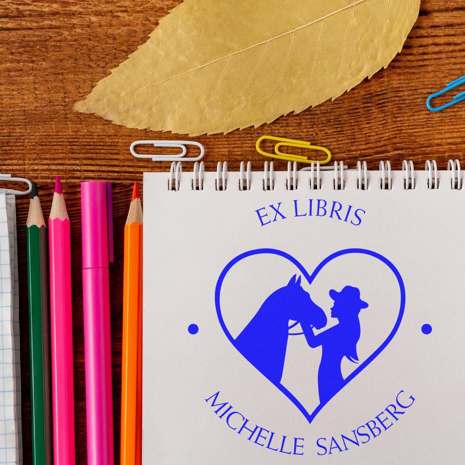 Blue equestrian heart ex libris book stamp and seal on white notebook, with colorful pencils, clips, and dried leaf.