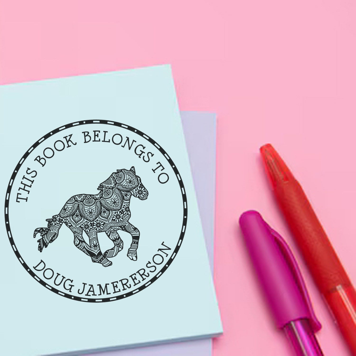 Light blue bookplate stamp seal with ornate horse and black text on pink background, beside red and purple pens.