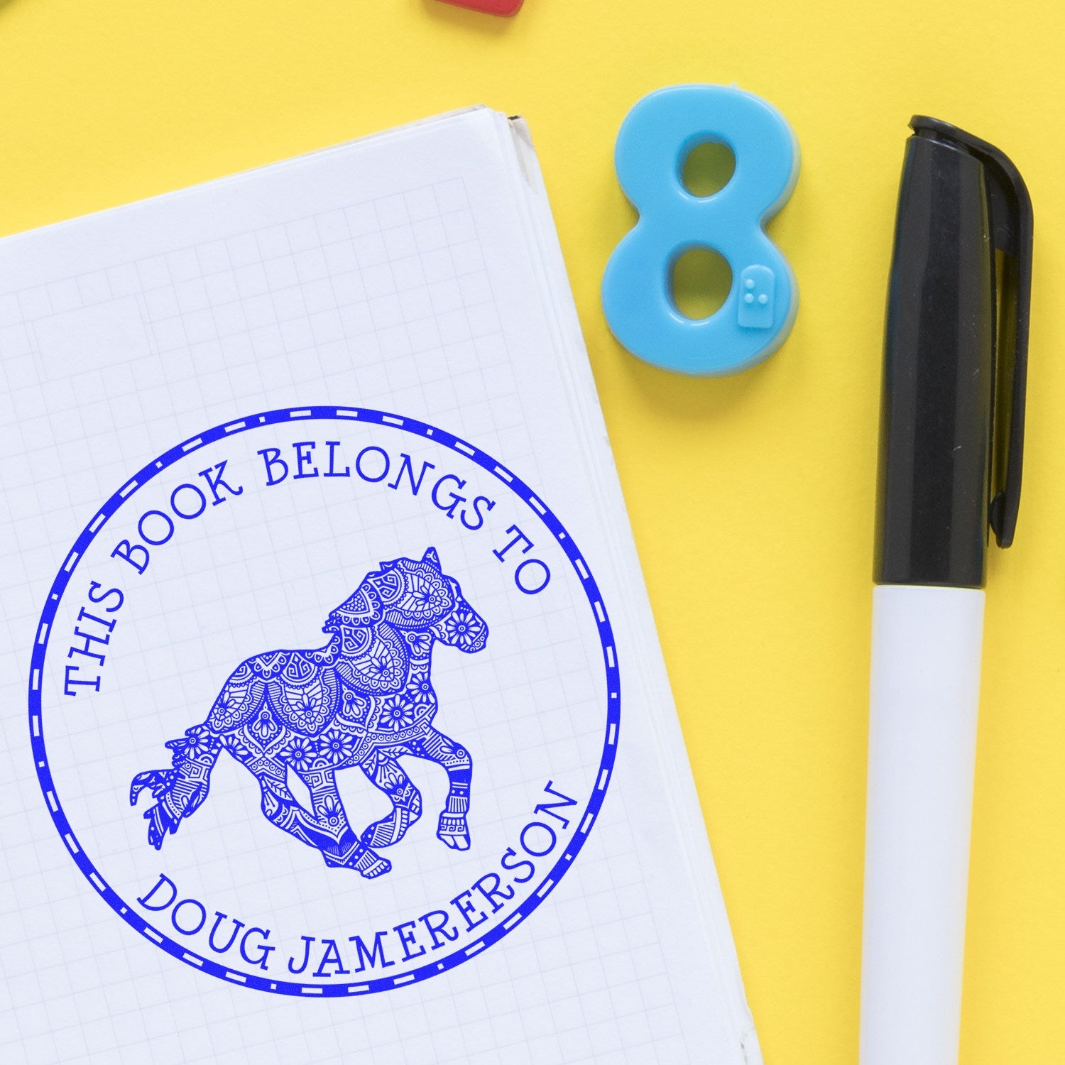 Blue custom book stamp and seal with horse design on white notebook, beside black marker on yellow background.