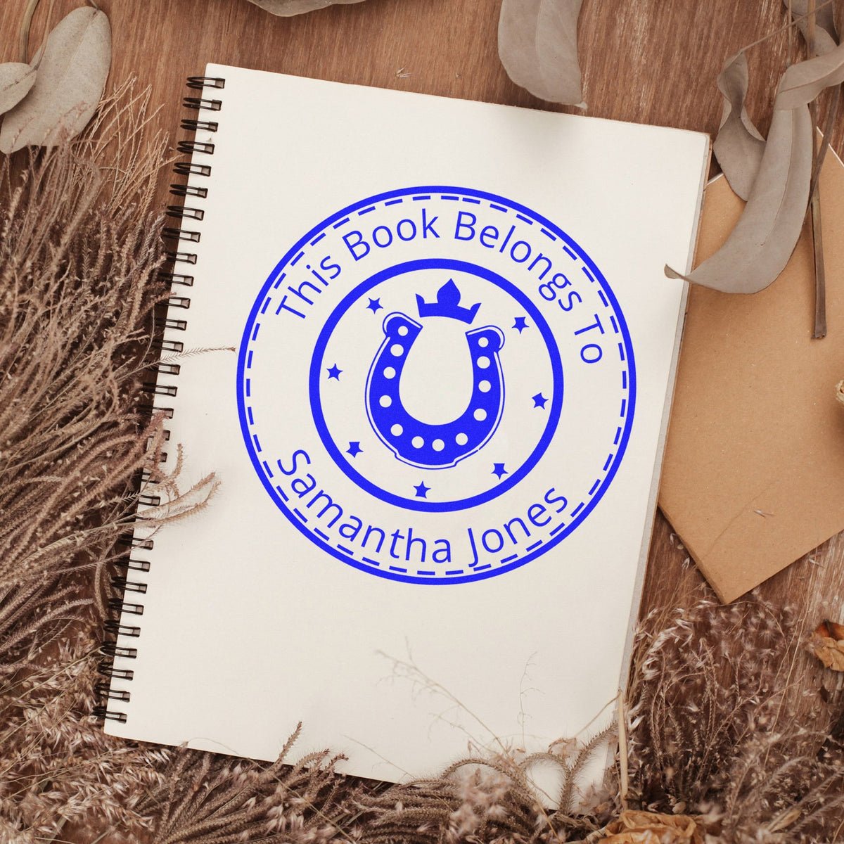 Blue personalized book stamp seal with horseshoe and stars on a cream spiral notebook, surrounded by dried florals.
