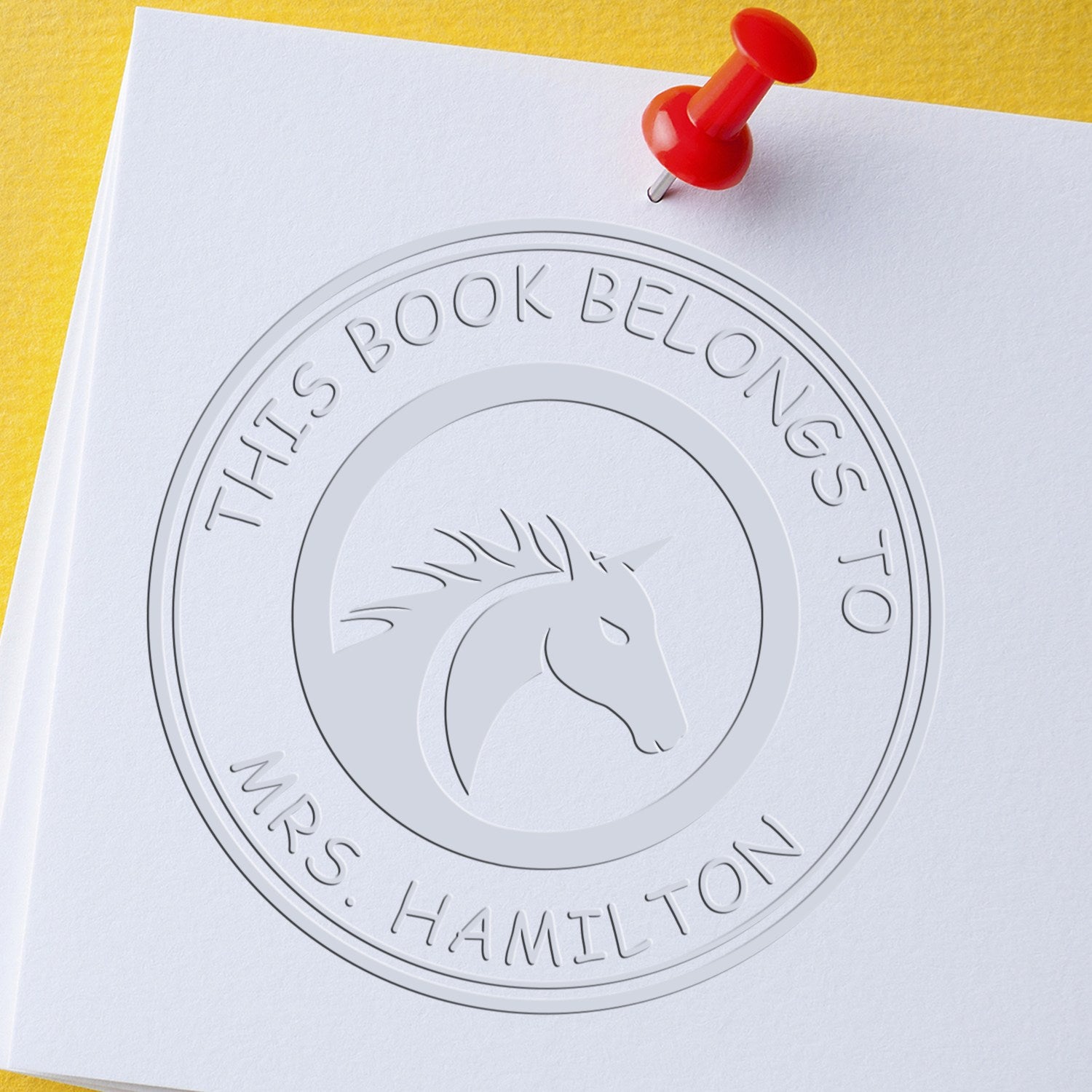 White embossed circular seal stamp with horse head logo and red pushpin on paper, on yellow background