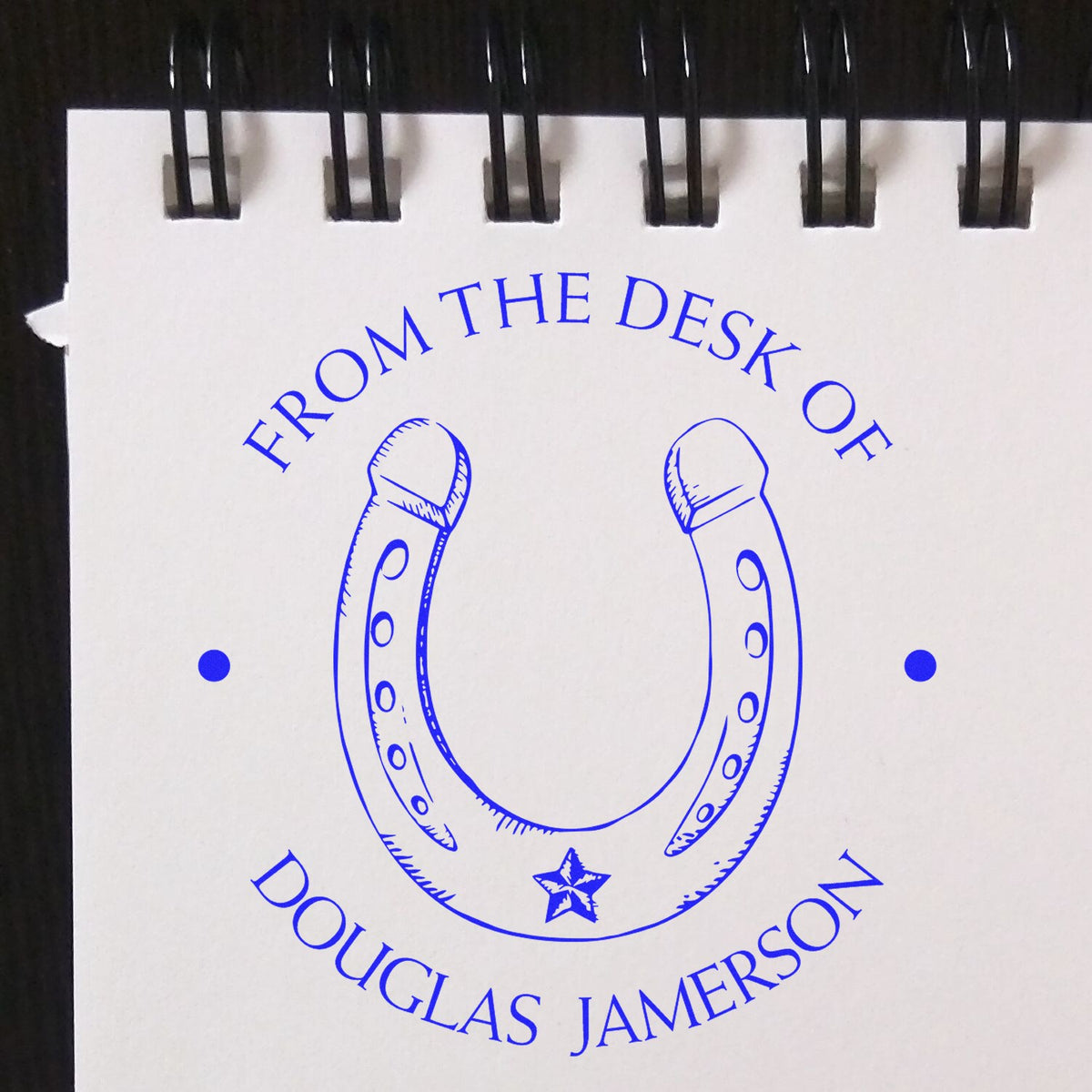 Blue horseshoe stamp seal in bright blue on white spiral notepad, reading From the Desk of Douglas Jamerson, with a small star.