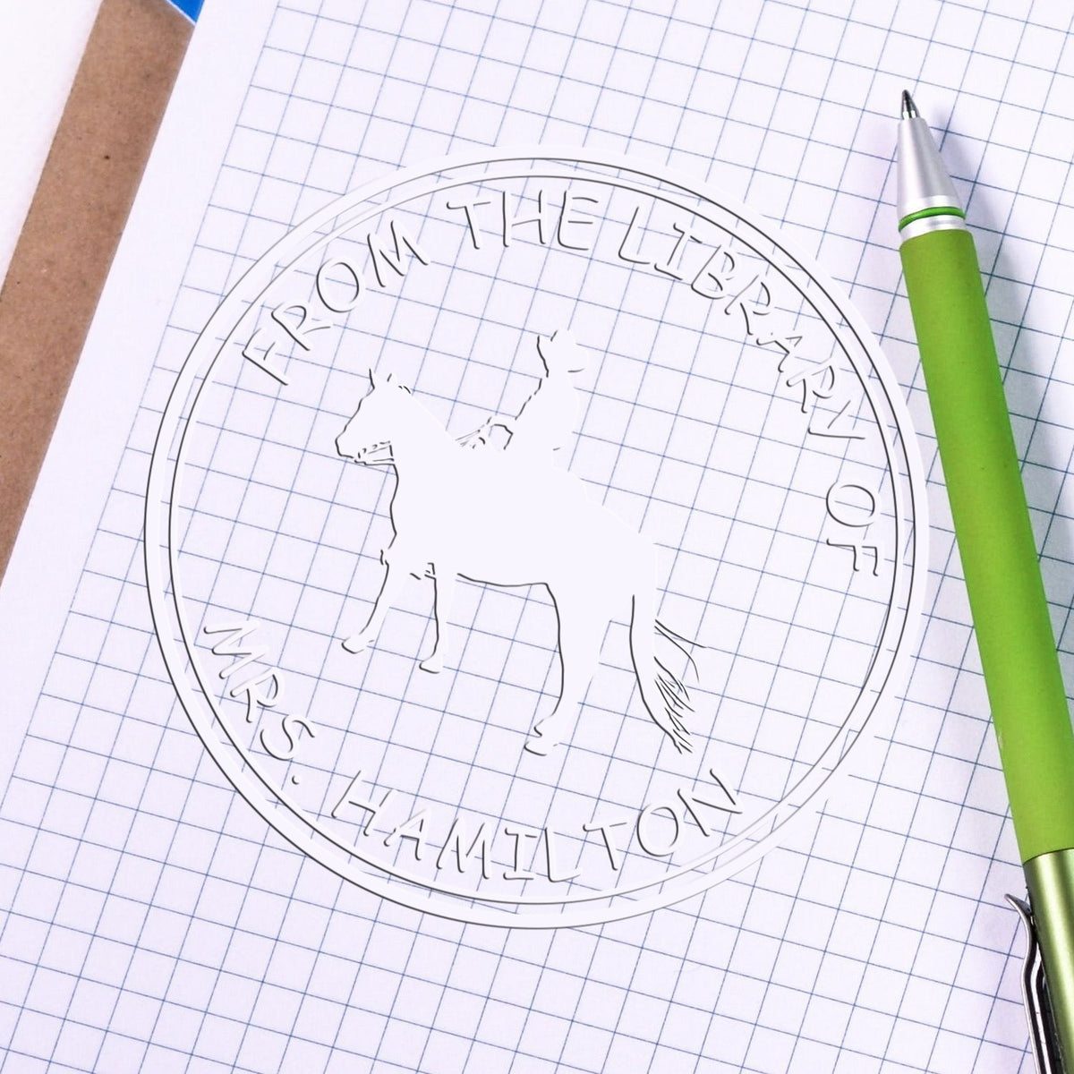 Transparent circular seal stamp with horse silhouette on graph paper, beside a green pen.