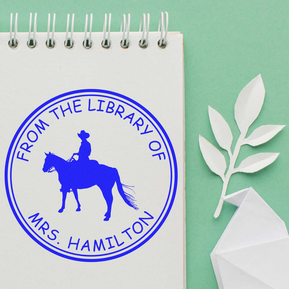 Blue round custom library stamp reading From the Library of Mrs. Hamilton on a white spiral notebook, with paper leaves on mint green.